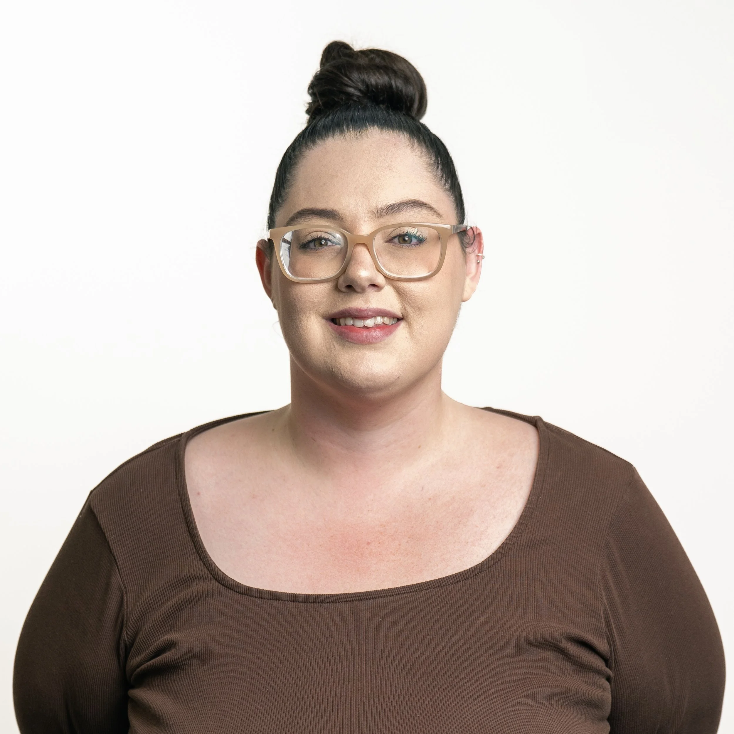 A woman with dark hair in a bun, wearing glasses and a brown shirt, smiling at the camera against a plain white background.