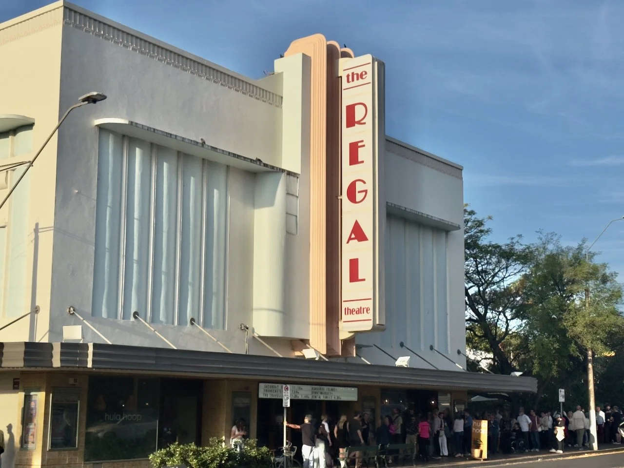 People lined up outside the Regent Theatre, a vintage-style theater building with a tall vertical sign that reads 'the REGAI' and 'theatre' at the bottom in red letters.