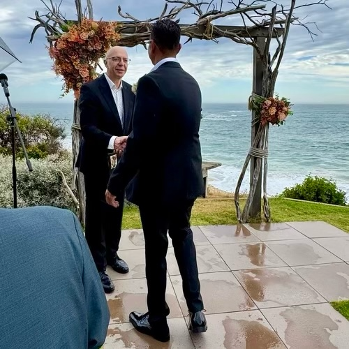 Two men in suits shaking hands during a wedding ceremony on an outdoor patio overlooking the ocean, with floral arrangements and a wooden arch.