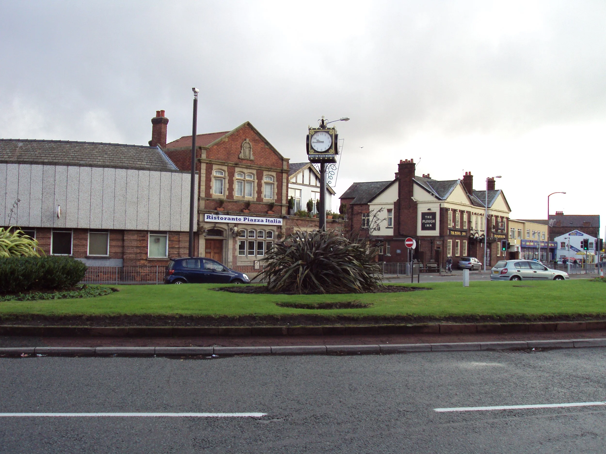 Street view with a roundabout, clock, and buildings including a restaurant, pub, and shops under cloudy sky.