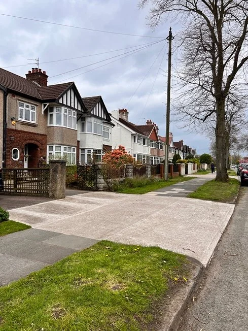 Residential street with row of houses, sidewalk, trees, and parked cars under cloudy sky.