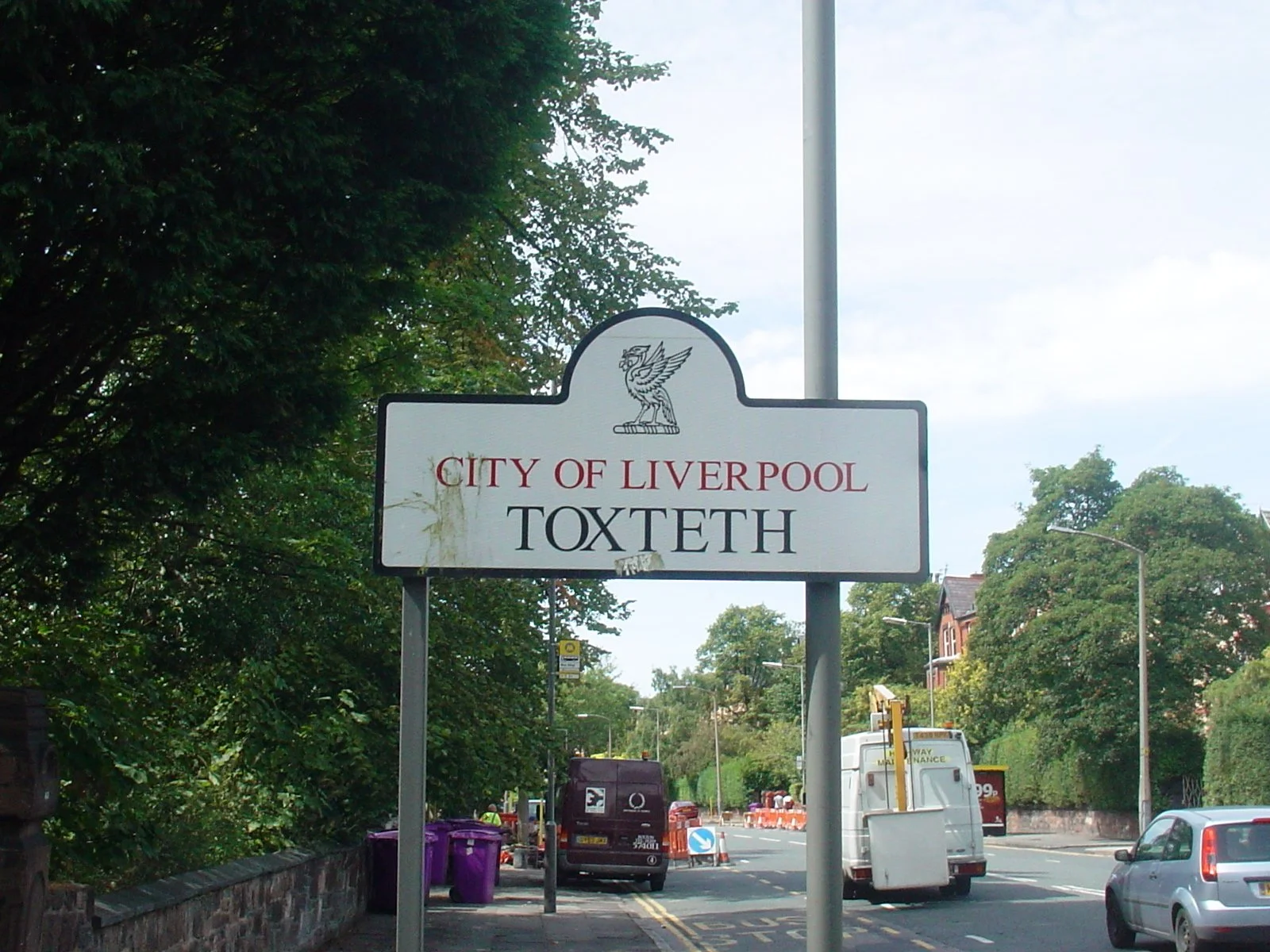 Street view with a sign reading 'City of Liverpool Toxteth,' trees on the left, parked cars, and a cloudy sky.