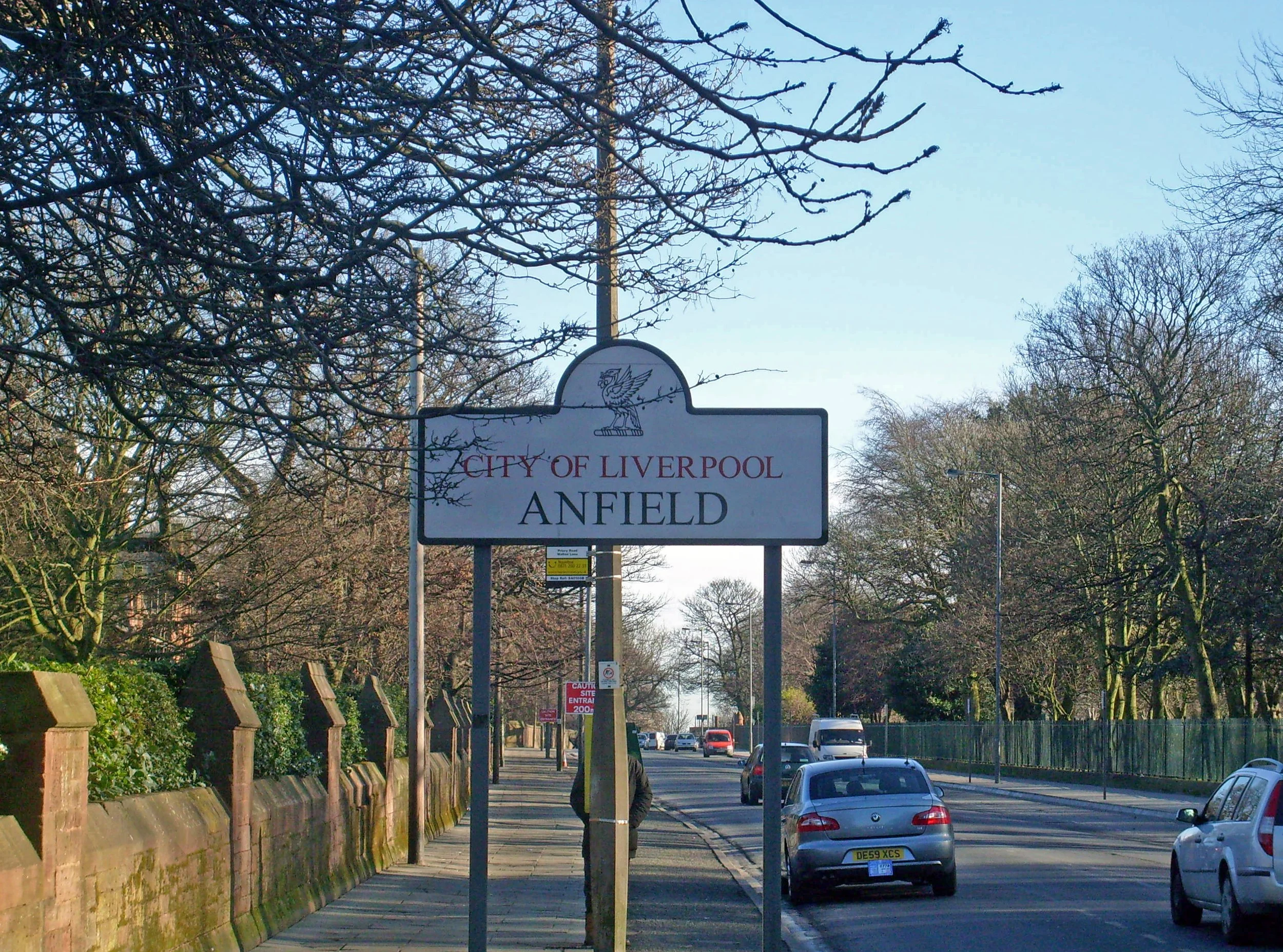 View of a street with cars parked and driving, trees on both sides, and a sign that reads 'City of Liverpool Anfield' with a bird illustration at the top.