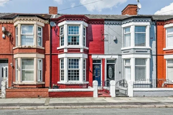 Row of colorful brick townhouses with bay windows, front steps, and small front yards, under a partly cloudy sky.