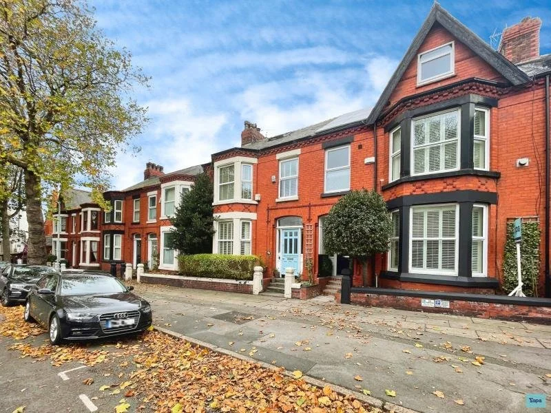 Row of Victorian-style brick houses with bay windows and small front gardens, parked cars on the street, and fallen autumn leaves.