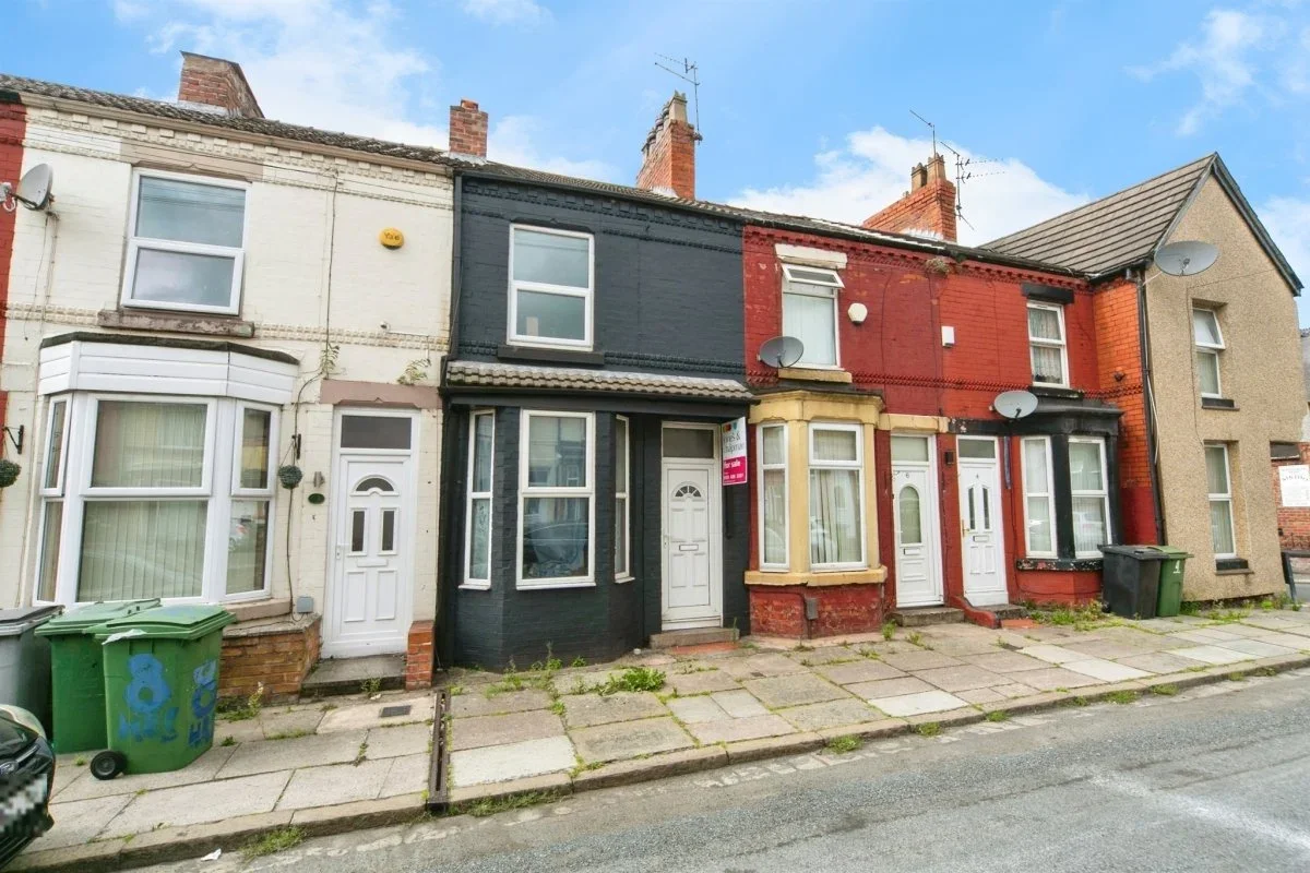 Row of attached classic houses with bay windows and brick facades, some painted in different colors, on a city street with scattered weeds, dumpsters, and satellite dishes.