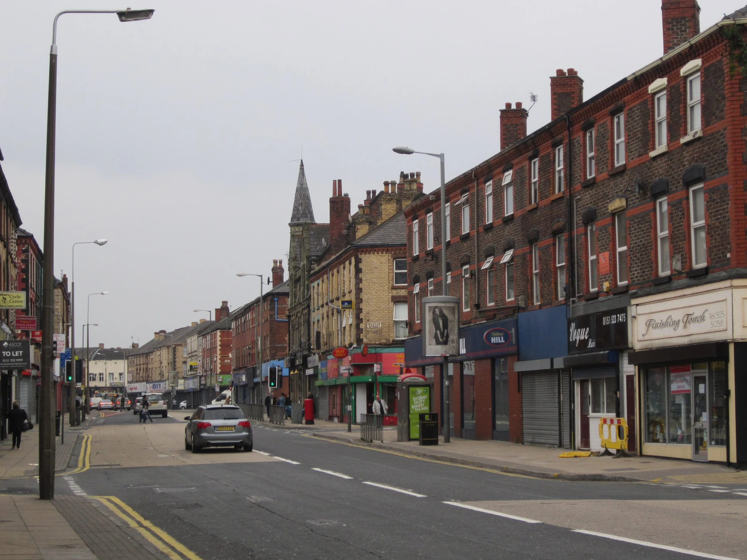 A city street with shops and businesses, parked cars, and a few pedestrians, under an overcast sky.