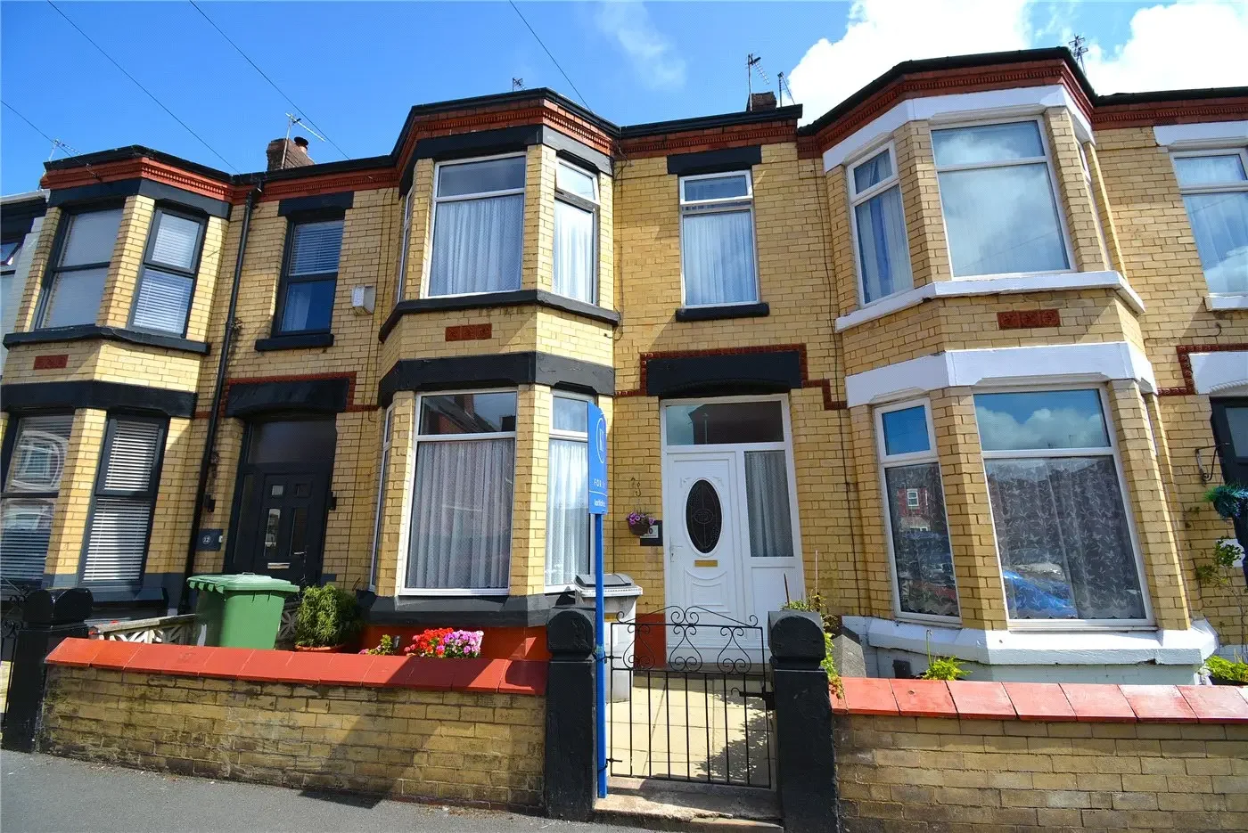 A row of yellow brick townhouses with black and white accents, bay windows, and small front gardens, under a blue sky with some clouds.