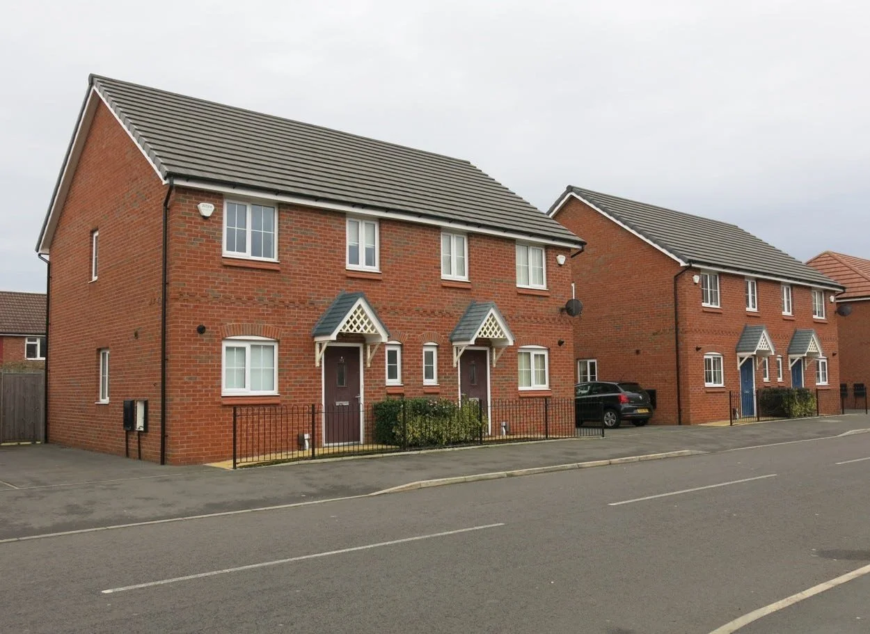 A row of modern red brick townhouses with small front yards and cars parked in driveways, under a cloudy sky.