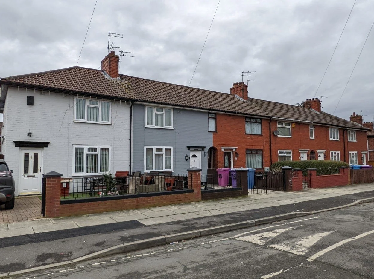 Row of attached houses with brick and painted facades, small front yards with fences, trash bins, and parked cars under a cloudy sky.