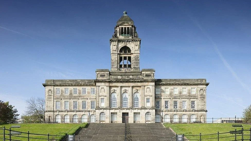A large, historic government building with a central clock tower, stone facade, and multiple windows, set on a grassy hill with stairs leading up to the entrance.