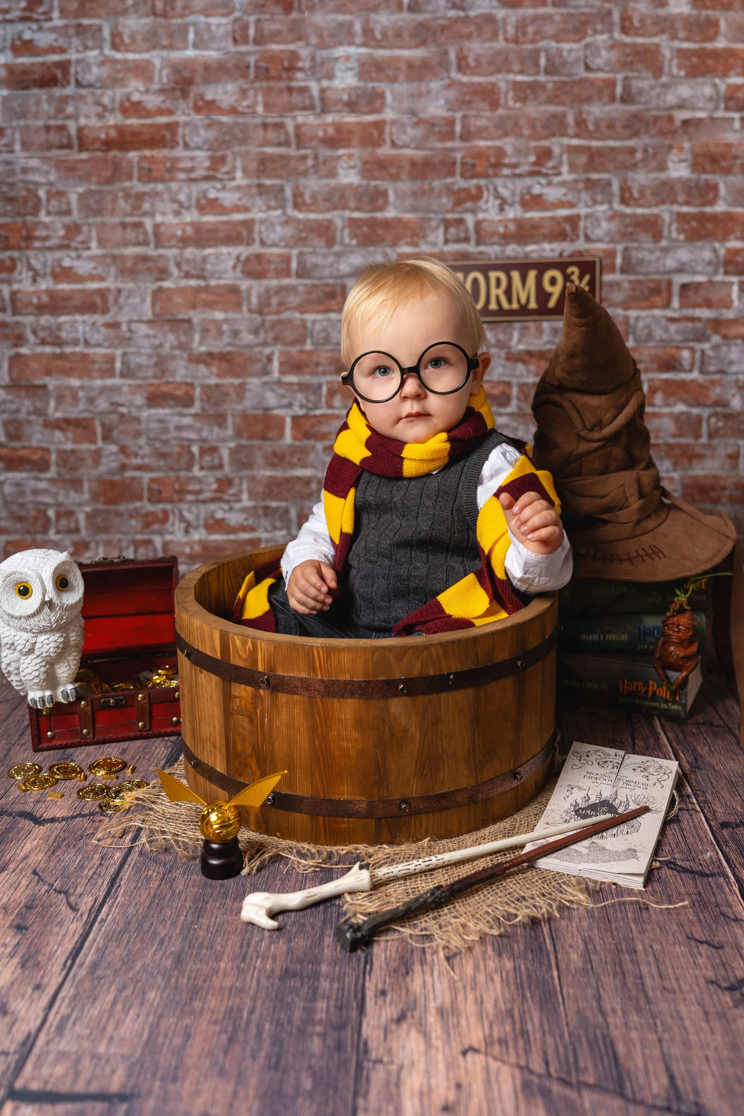 Kleiner Junge in Harry-Potter-Kostüm mit Brille, Schal, in einer Holzkiste, umgeben von Harry-Potter-Accessoires, einem Zauberstab, Büchern, einer Eule, einem Zauberhut, einem Brief und Goldmünzen vor einer Backsteinwand im Zimmer.
