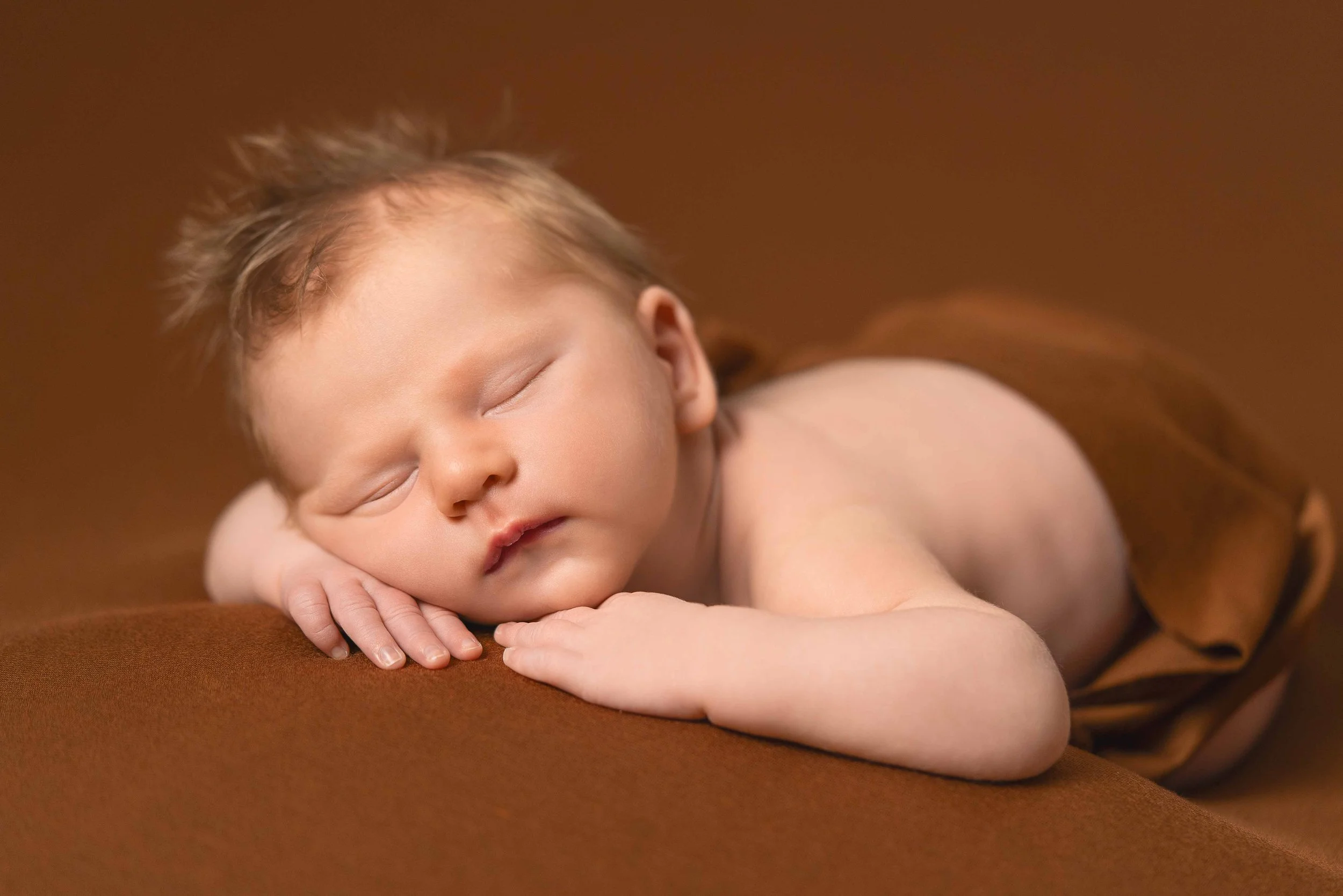 Ein schlafender Säugling liegt auf einem braunen Stoffhintergrund, mit dem Kopf auf den Händen ruhend.
