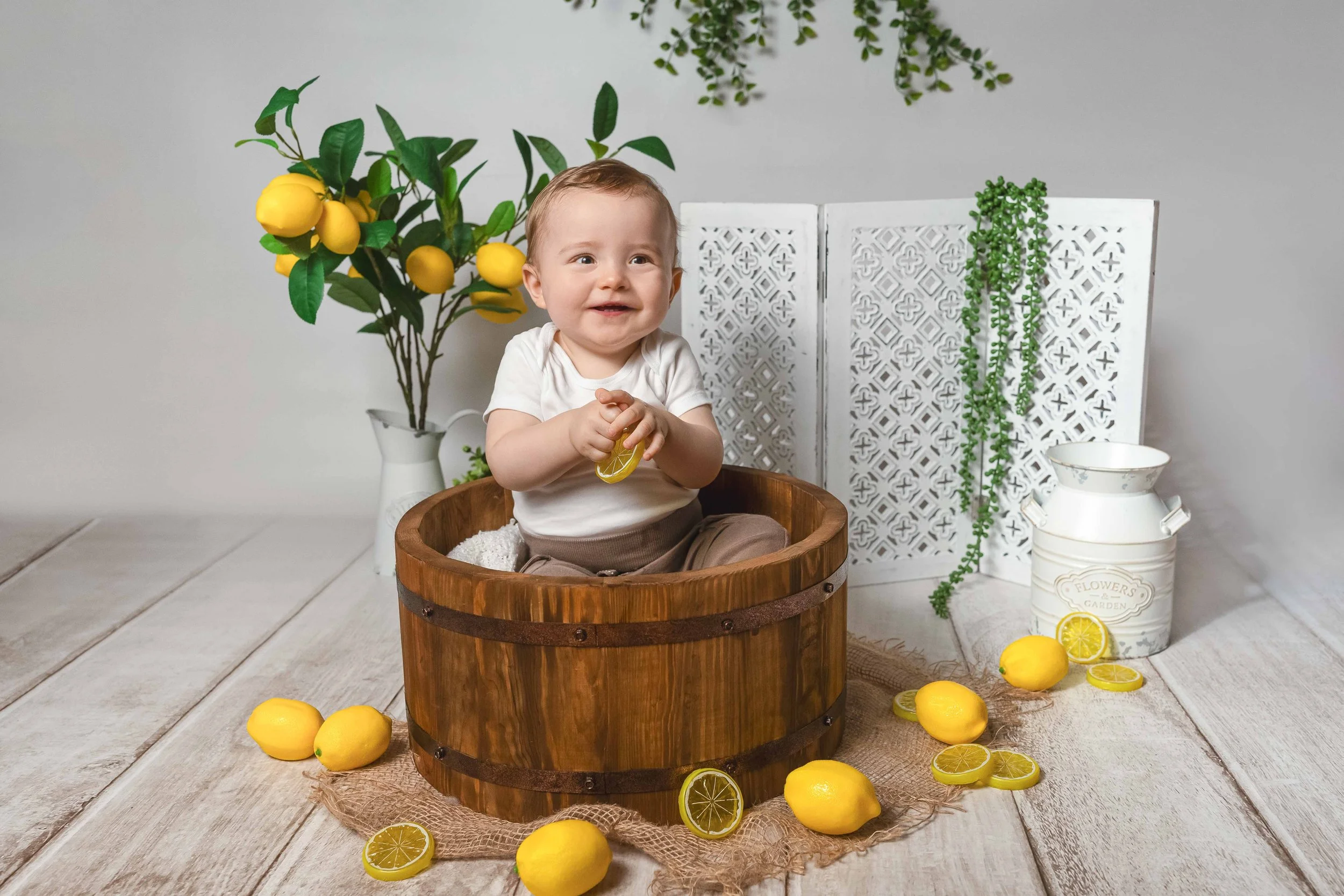 Lachender Baby in einem runden Holzkorb, umgeben von Zitronen, im Hintergrund Zitronenbaum, weiße Dekoration, Papierschirm, weißer Blumentopf mit Gärten, Holzfußboden.