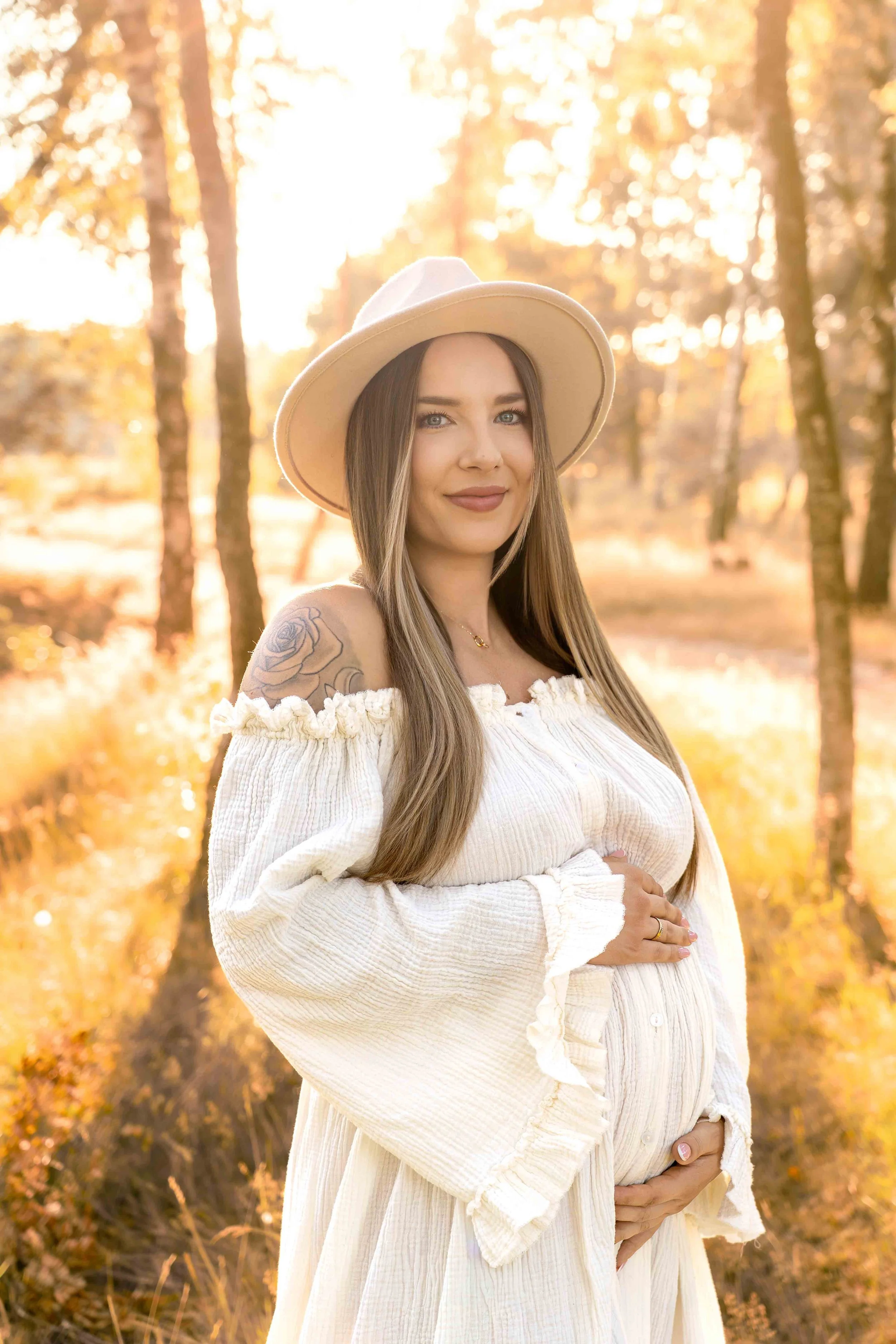 Eine schwangere Frau mit langen Haaren, die einen weiten, weißen, lockeren Rock und ein passendes Oberteil trägt, in einer herbstlichen Waldumgebung mit goldenen Blättern, Sonnenlicht und Bäumen im Hintergrund.