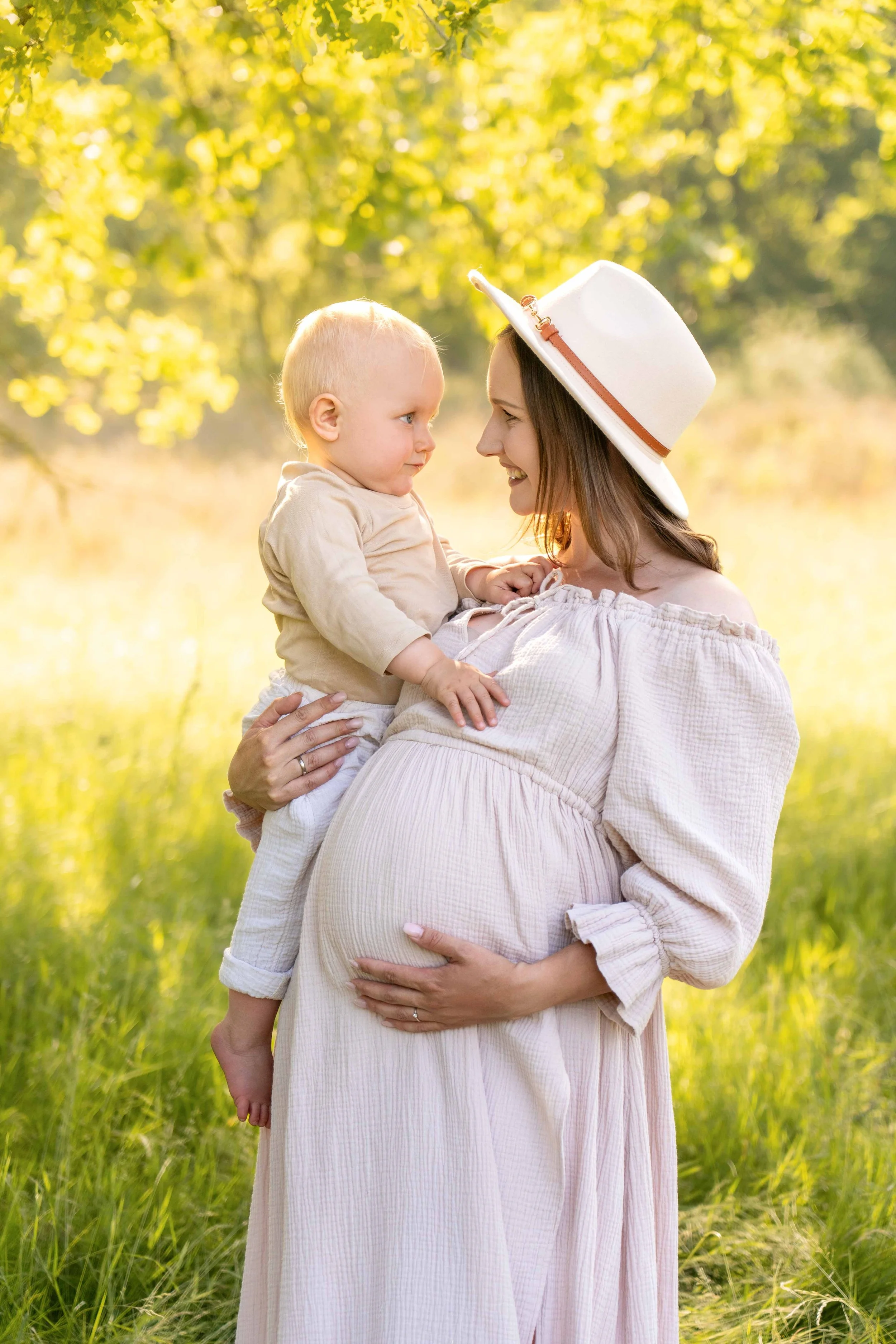 Eine schwangere Frau hält ein kleines Kind in einem sonnigen, grünen Park.