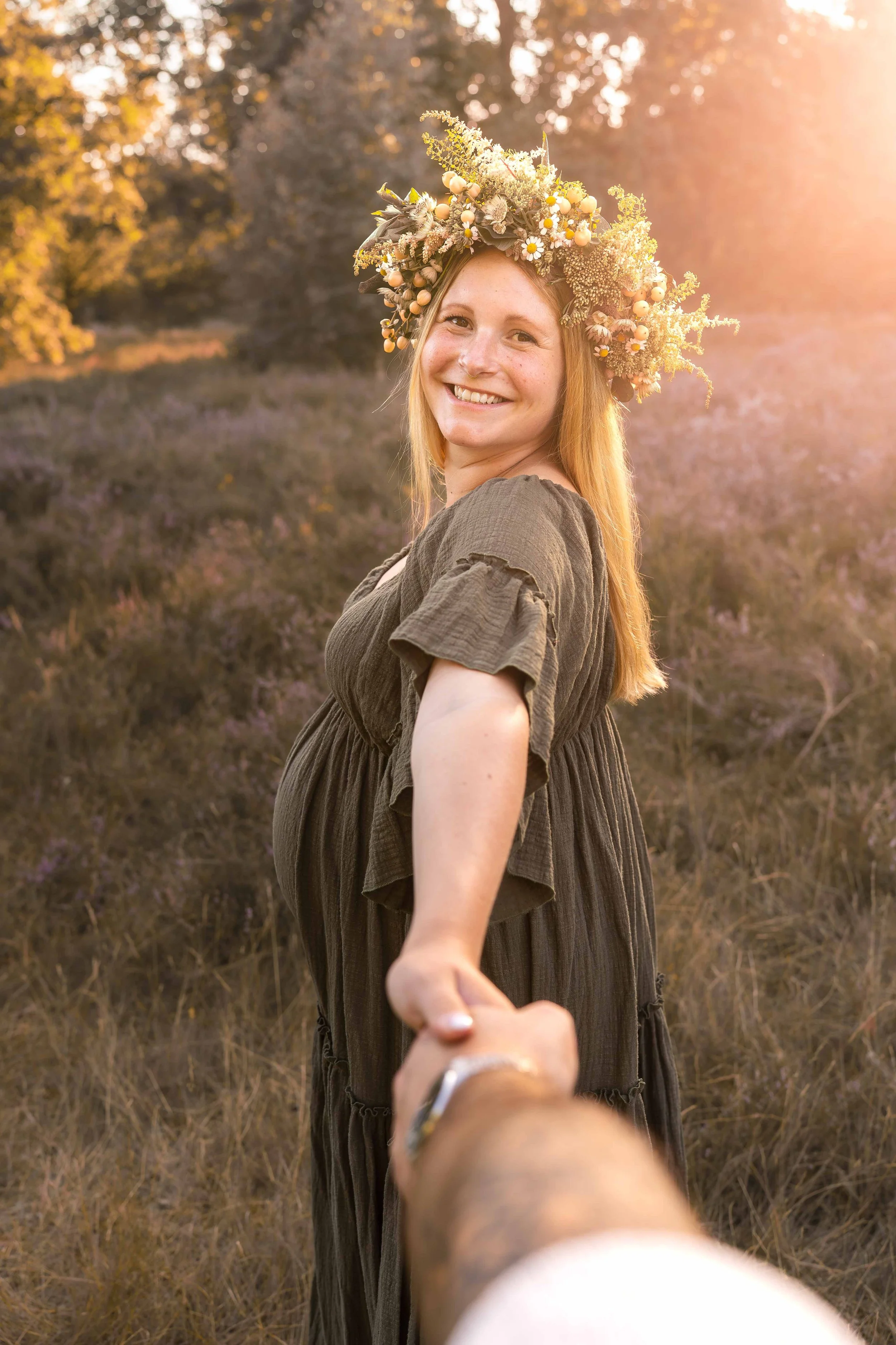 Junge Frau mit Blumenkranz im Haar, die lächelnd eine Hand hält, im Sonnenuntergang in einem Feld mit Büschen und Bäumen im Hintergrund.