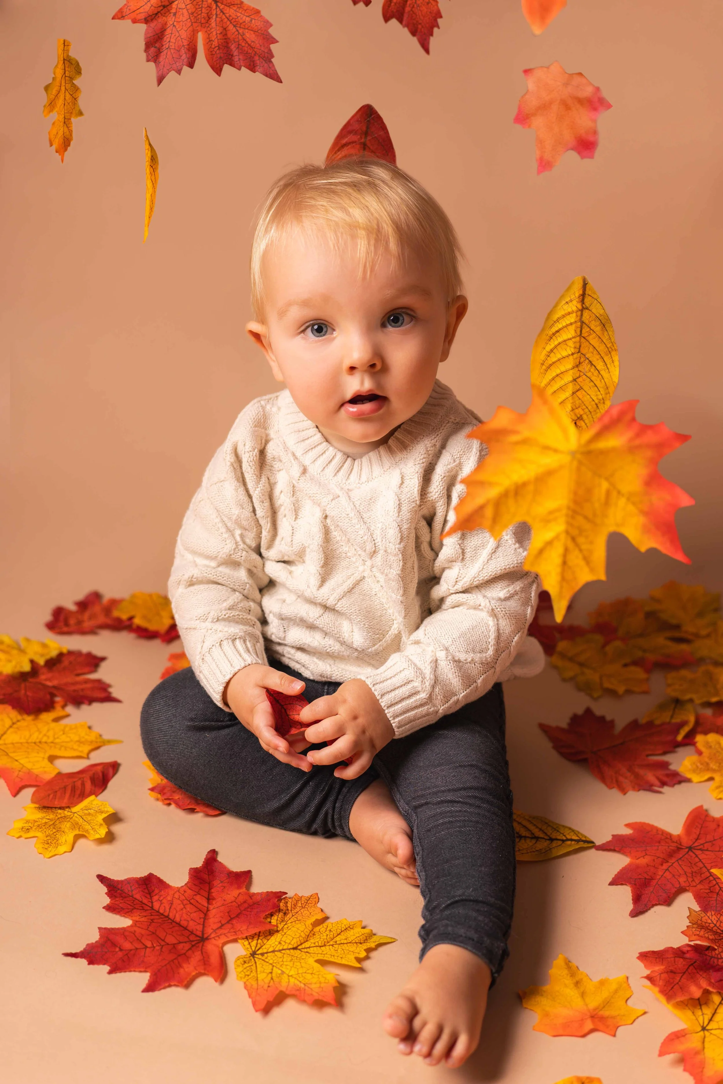 Ein Baby sitzt auf einem beige Hintergrund, umgeben von herabfallenden Herbstblättern in Rot, Orange und Gelb. Das Baby trägt einen cremefarbenen Pullover und dunkle Hosen, hält ein rotes Herbstblatt in der Hand und schaut neugierig in die Kamera.