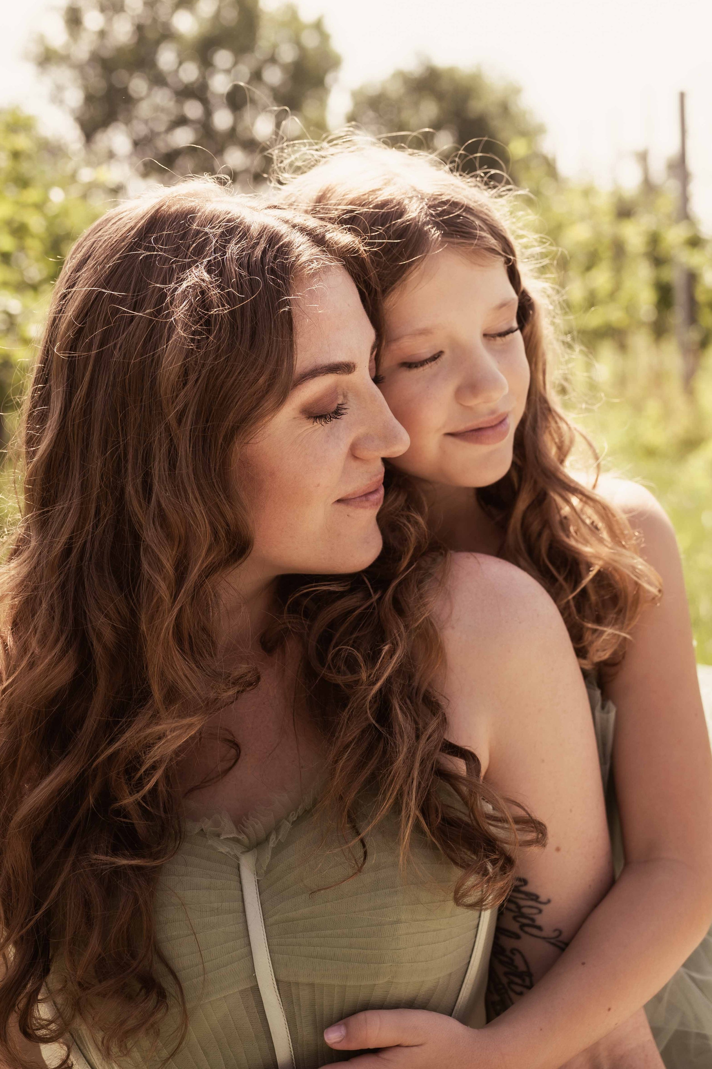 Eine Frau und ein Mädchen, die eng aneinander kuscheln, beide mit geschlossenen Augen und einem friedlichen Ausdruck im Gesicht, draußen bei Sonnenlicht, umgeben von grünen Pflanzen und Bäumen.