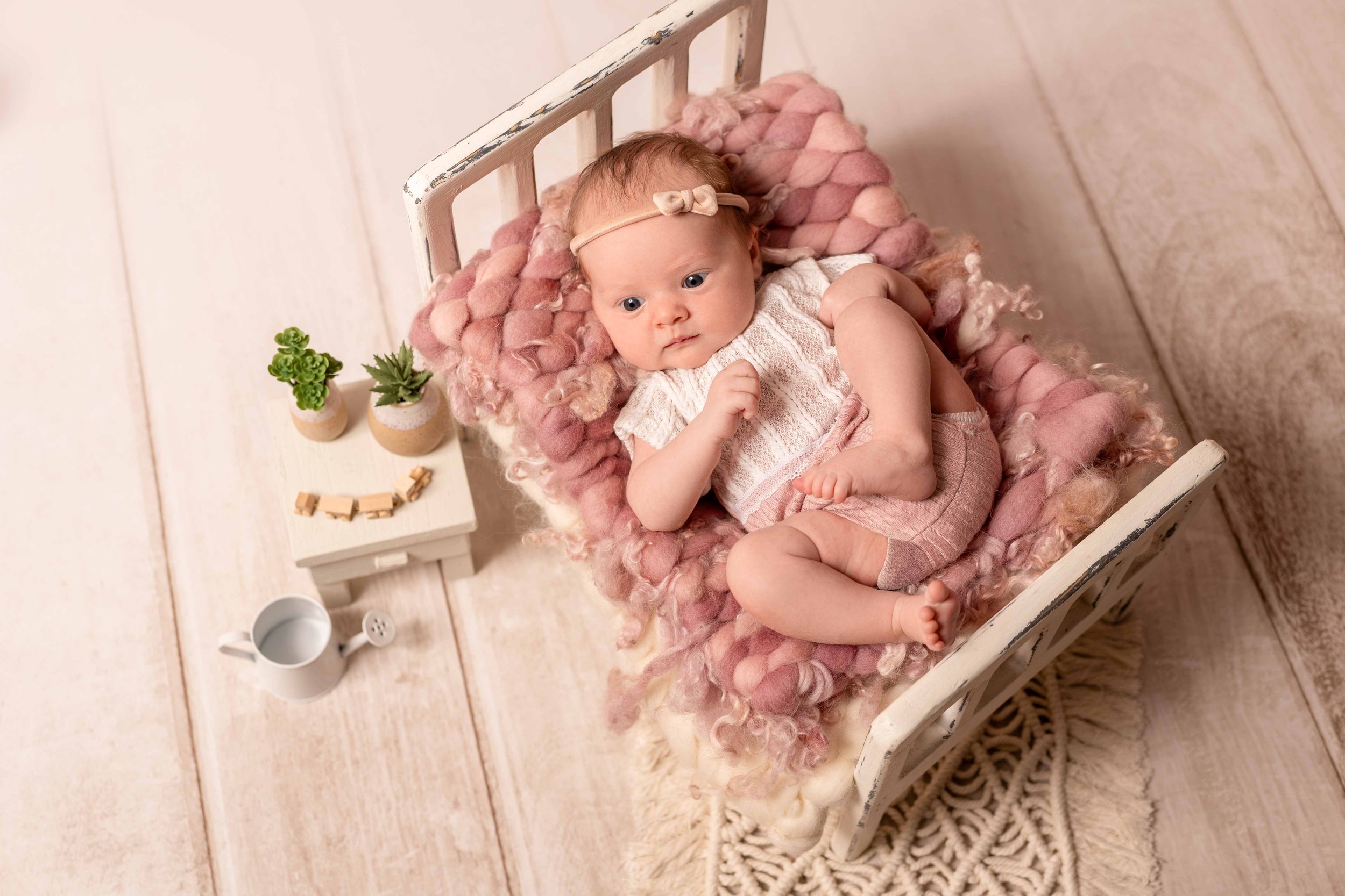 Süßes Baby liegt in einem kleinen weißen Kinderbett auf einer Pinken, kuscheligen Decke, umgeben von kleinen Pflanzen, einem weißen Milk-Can mit Wasser und einem weißen Nachttisch mit Holzklammern auf einem hellen Holzboden.