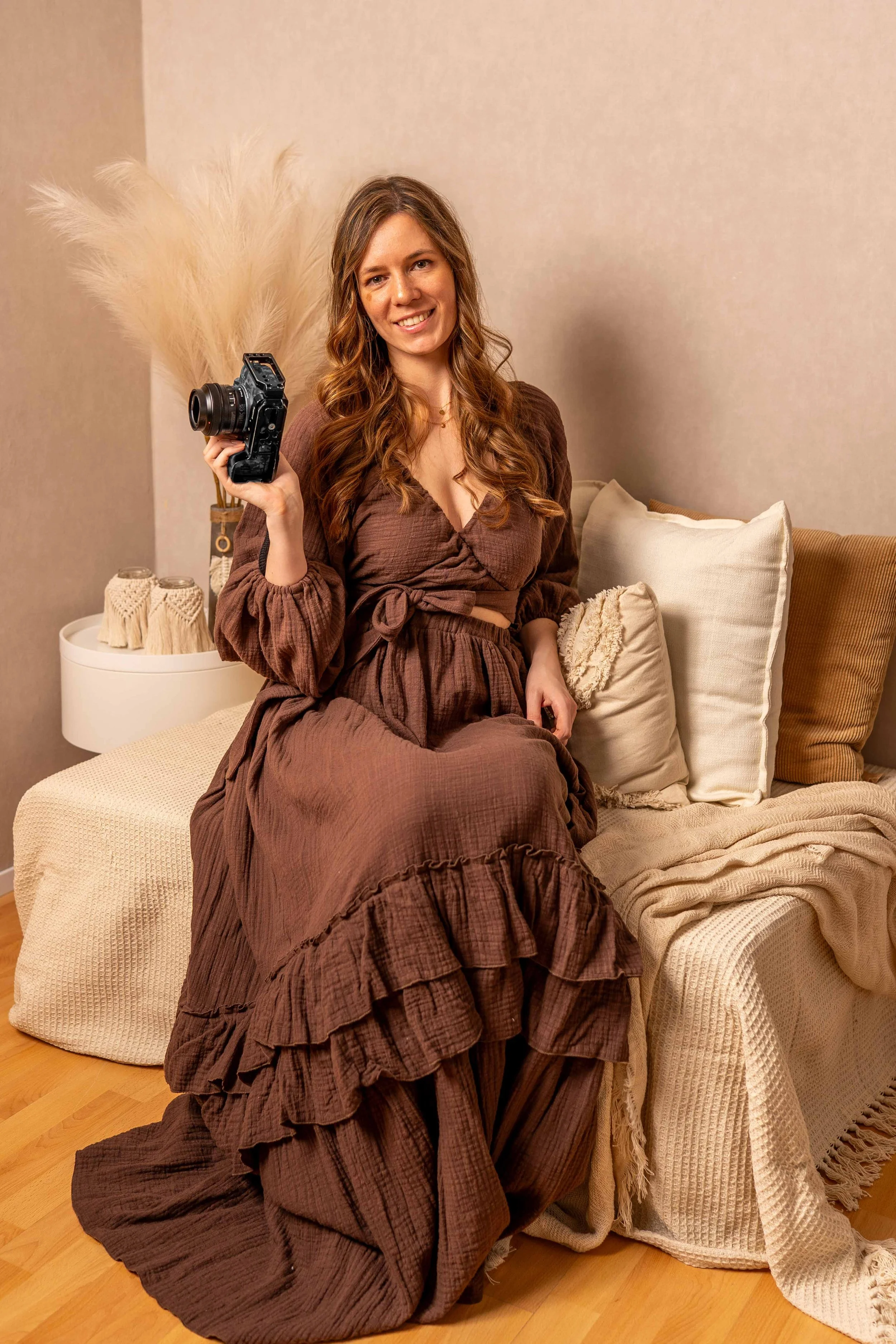 Eine junge Frau mit langen braunen Haaren sitzt auf einem Bett, hält eine Kamera in der Hand und lächelt.