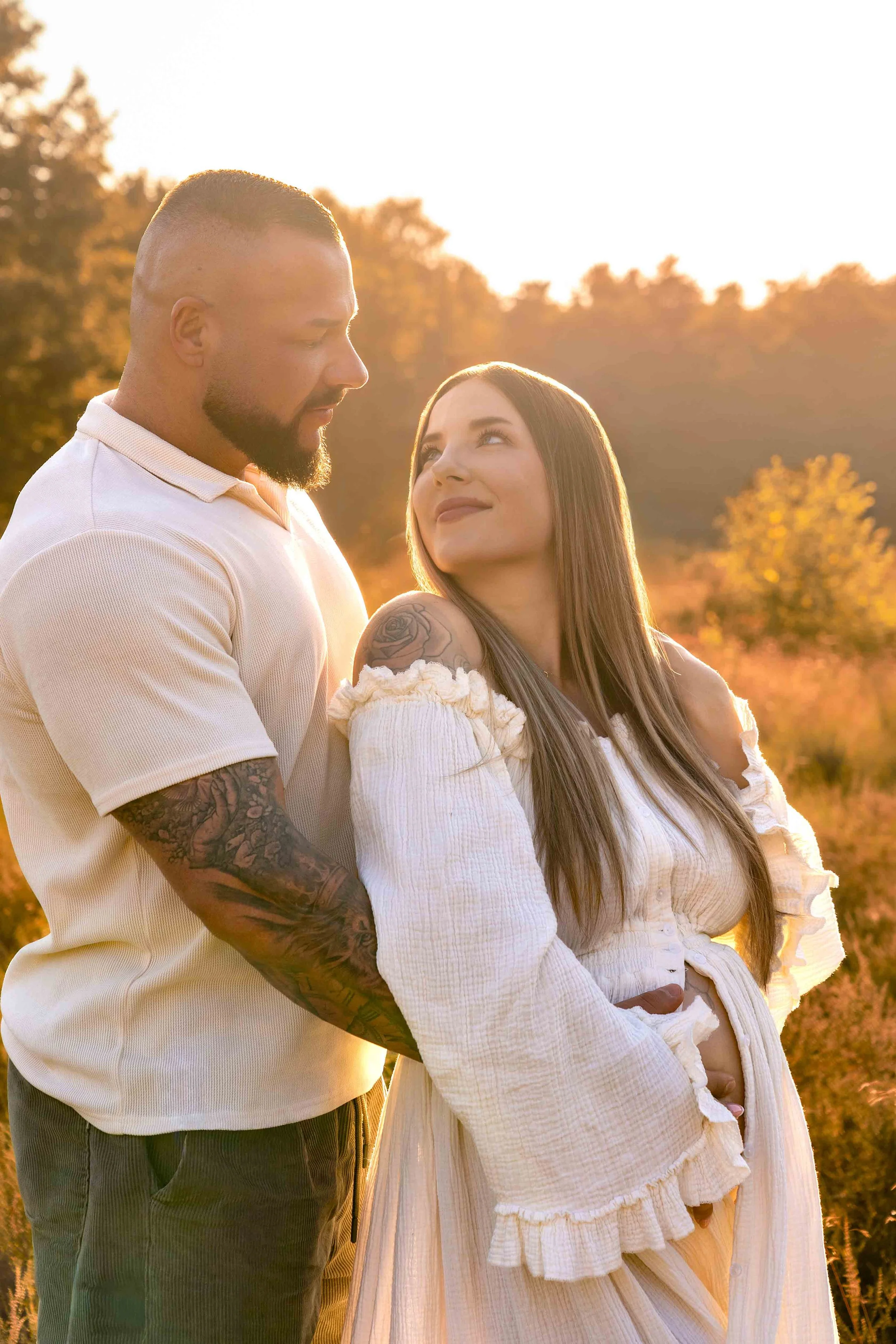 Ein Mann mit Tattoos hält eine Frau im weißen Kleid im Sonnenuntergang umarmt, beide schauen sich liebevoll an.