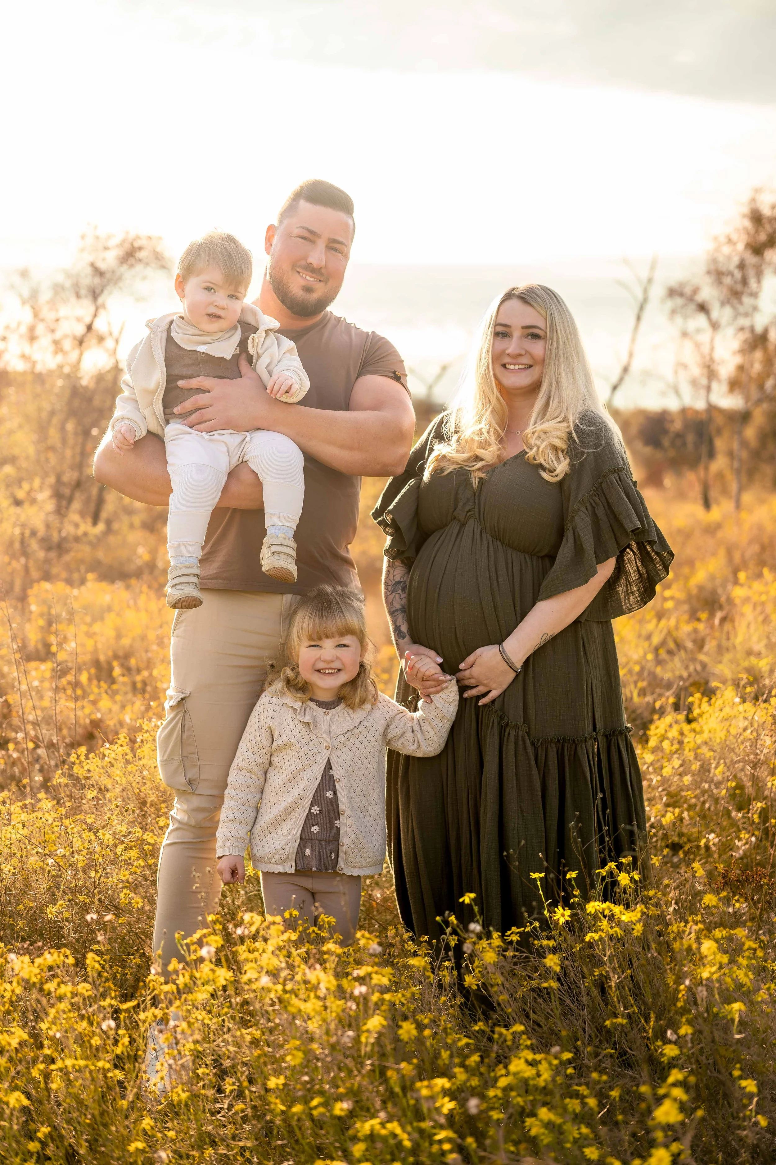 Familie im Sonnenuntergang in einem blühenden Feld, bestehend aus Vater, Mutter, einem kleinen Mädchen und einem Baby, alle lächelnd.