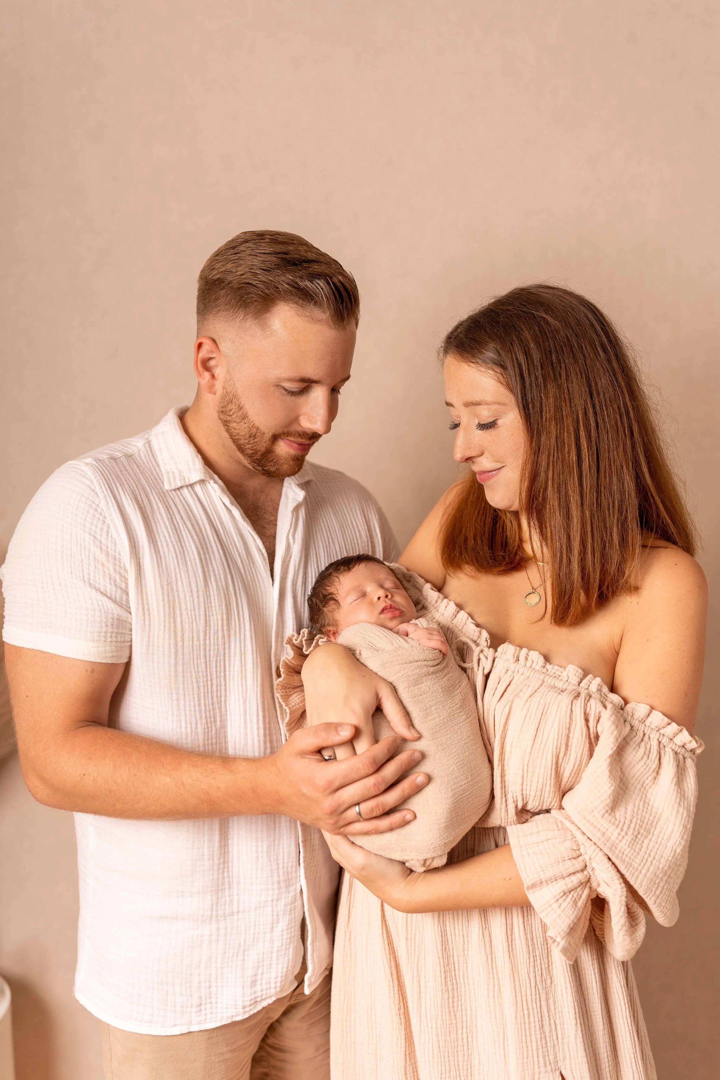 Ein Paar hält ihr neugeborenes Baby in den Armen, während sie liebevoll blicken, vor einem neutralen Hintergrund.