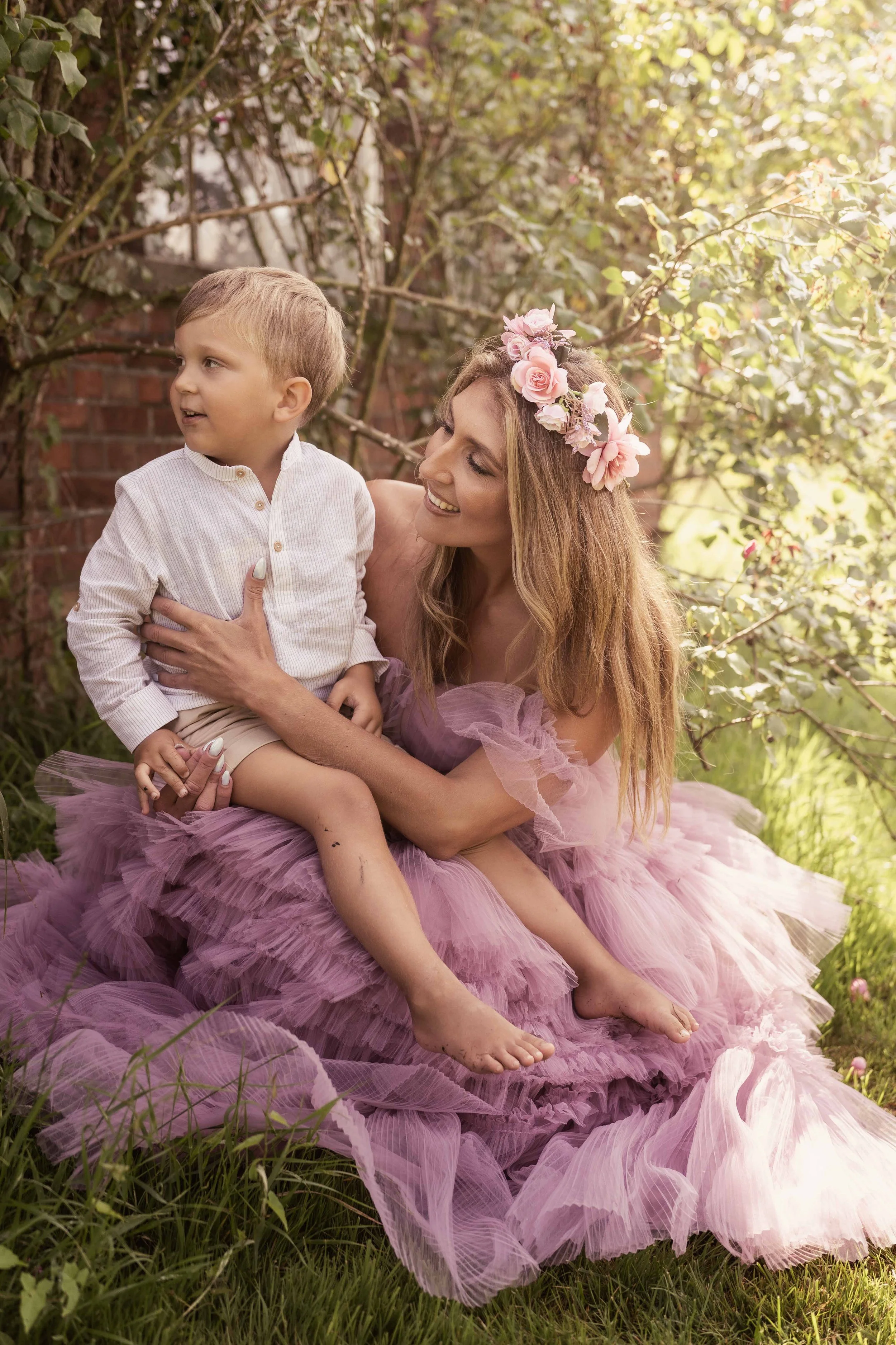 Eine lachende Frau mit Blumenkranz im Haar, die ein junges Kind auf ihrem Schoß hält, im Garten sitzend auf einem pinken Tüllkleid, umgeben von Büschen und Sonnenlicht.