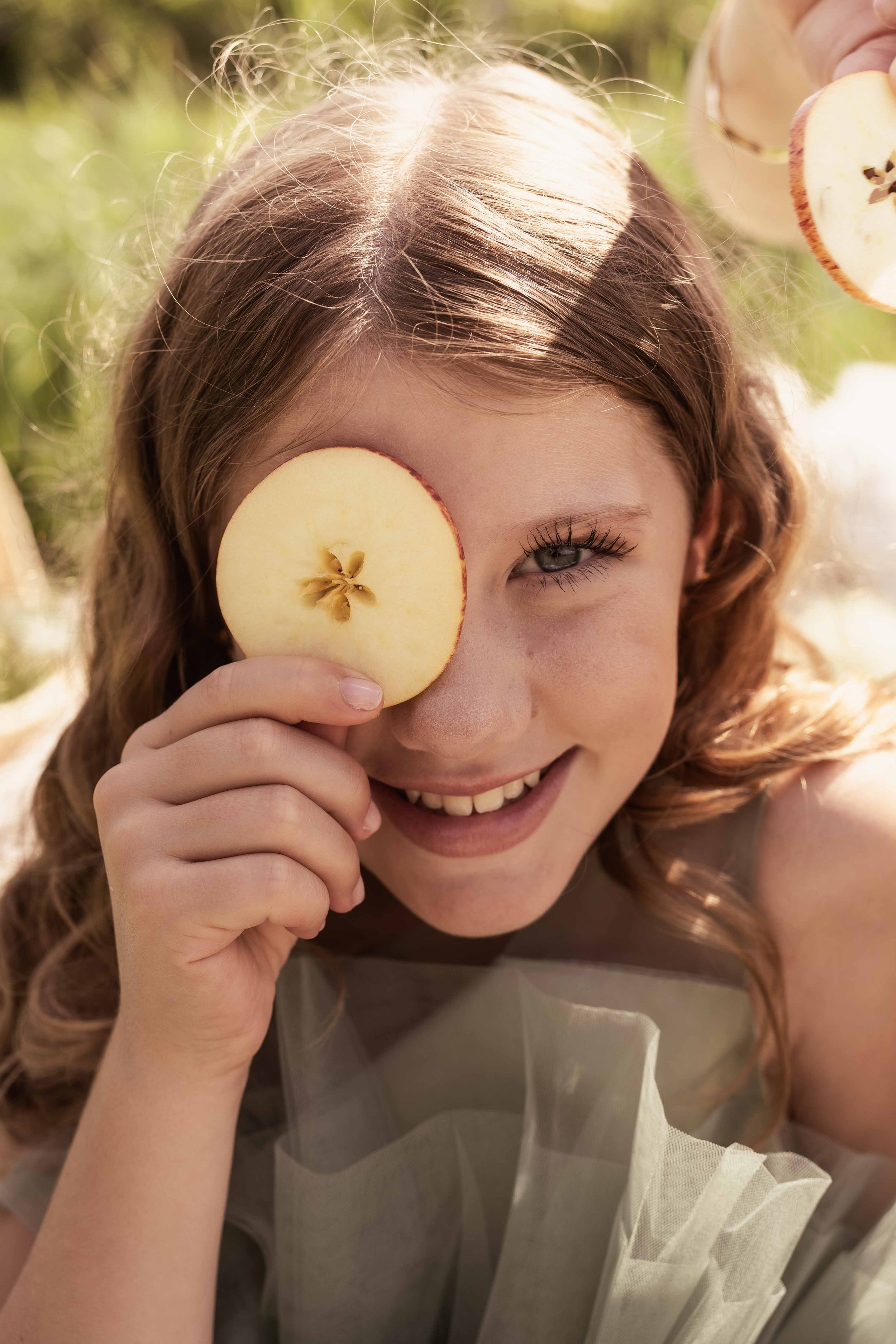 Ein junges Mädchen hält eine Apfelscheibe vor ihr rechtes Auge und lächelt in die Kamera, im Hintergrund ist grüne Natur sichtbar.