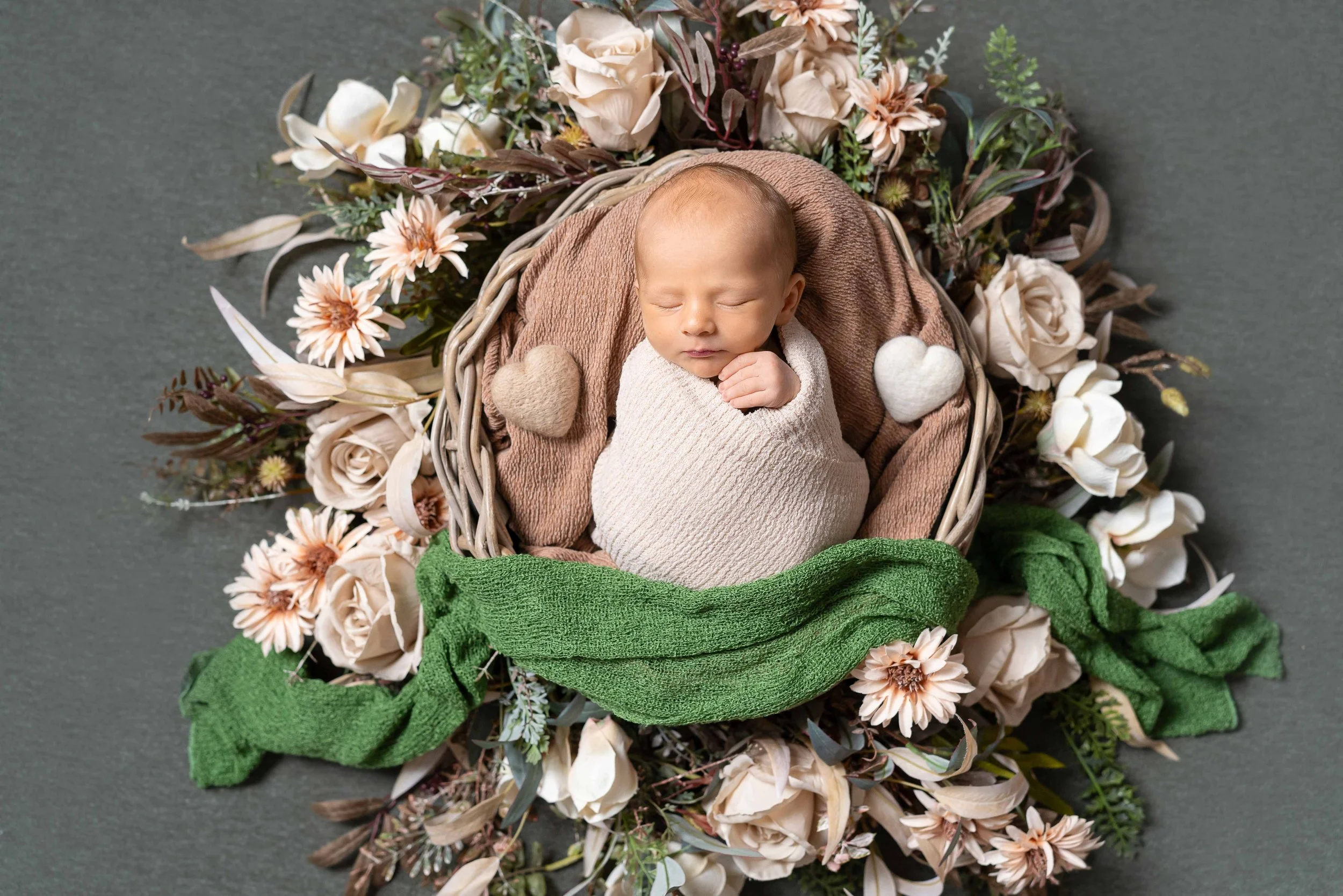 Ein schlafender neugeborener Baby in einem Korb, umgeben von Blumen und einem grünen Tuch, mit zwei Stoffherzen neben ihm.