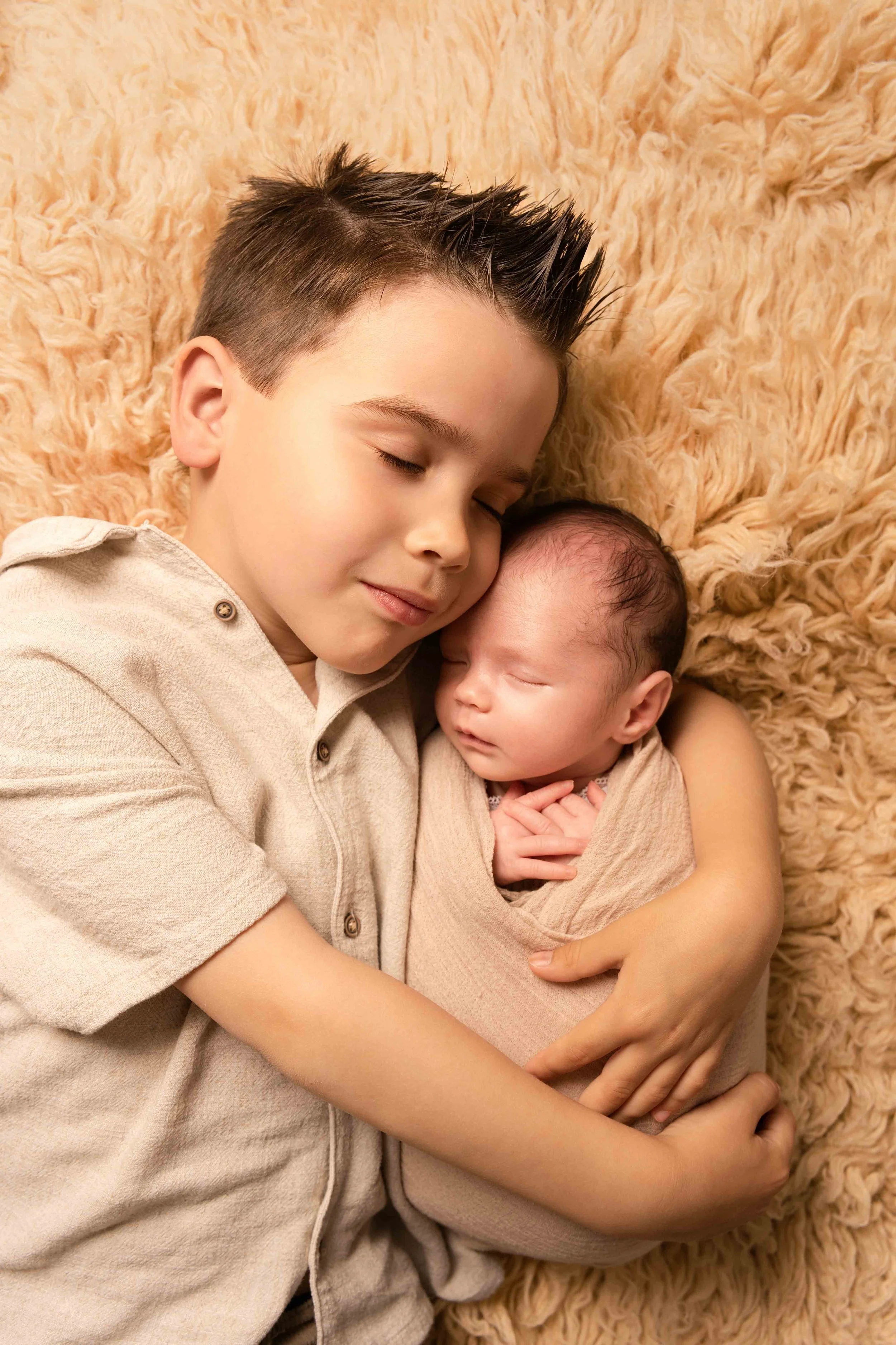 Ein älterer Junge mit braunem, gestyltem Haar hält ein schlafendes Baby in seinen Armen, beide liegen auf einem weichen, beigefarbenen Fell.