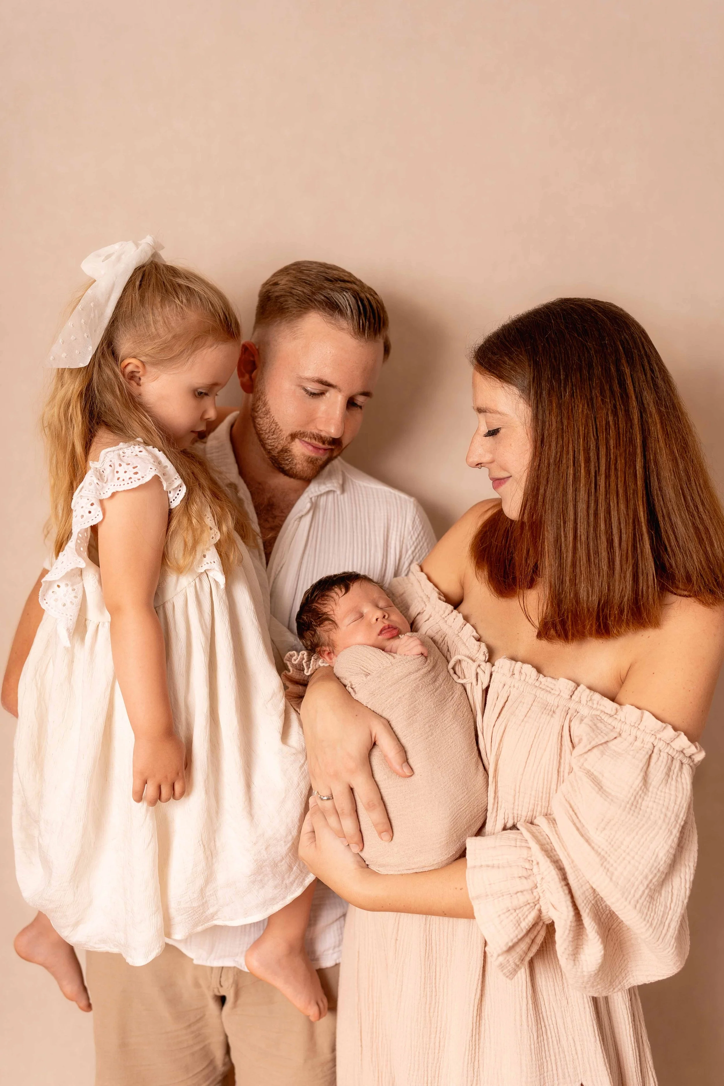 Familie mit zwei Erwachsenen und zwei Kindern, eines Babys und eines schulpflichtigen Mädchens, porträtiert in einem beige Hintergrund, Mom hält das Baby im Arm, alle schauen liebevoll auf das Baby