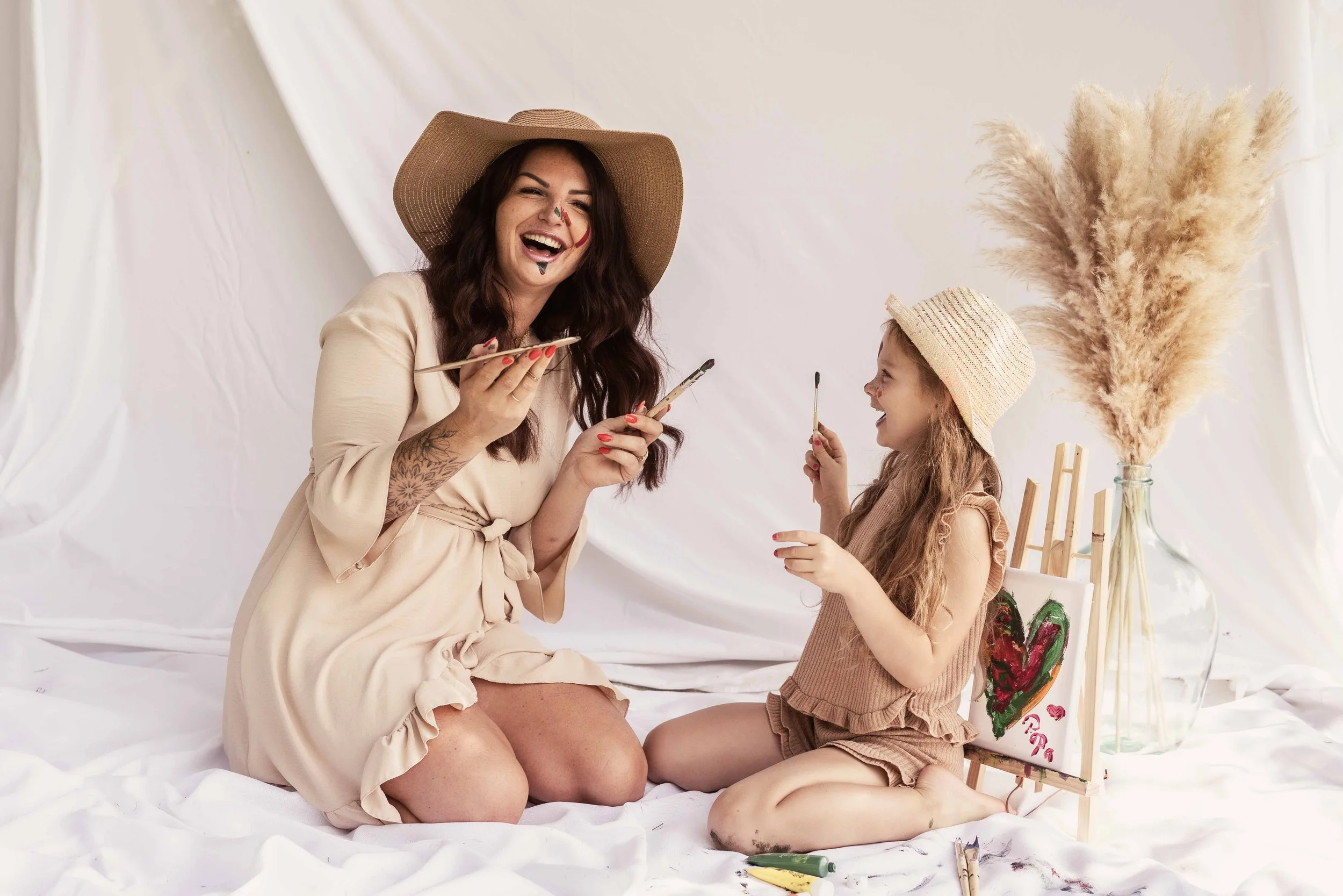 Eine lachende Frau mit einem Strohhut und einer tätowierten Hand malt mit einem Pinsel, während ein lachendes Mädchen mit Strohhut neben ihr sitzt und malt. Sie sind in einem hellen Raum mit weißen Vorhängen. Es gibt eine Vase mit getrocknetem Pampasgras und eine kleine Staffelei mit einem Gemälde.