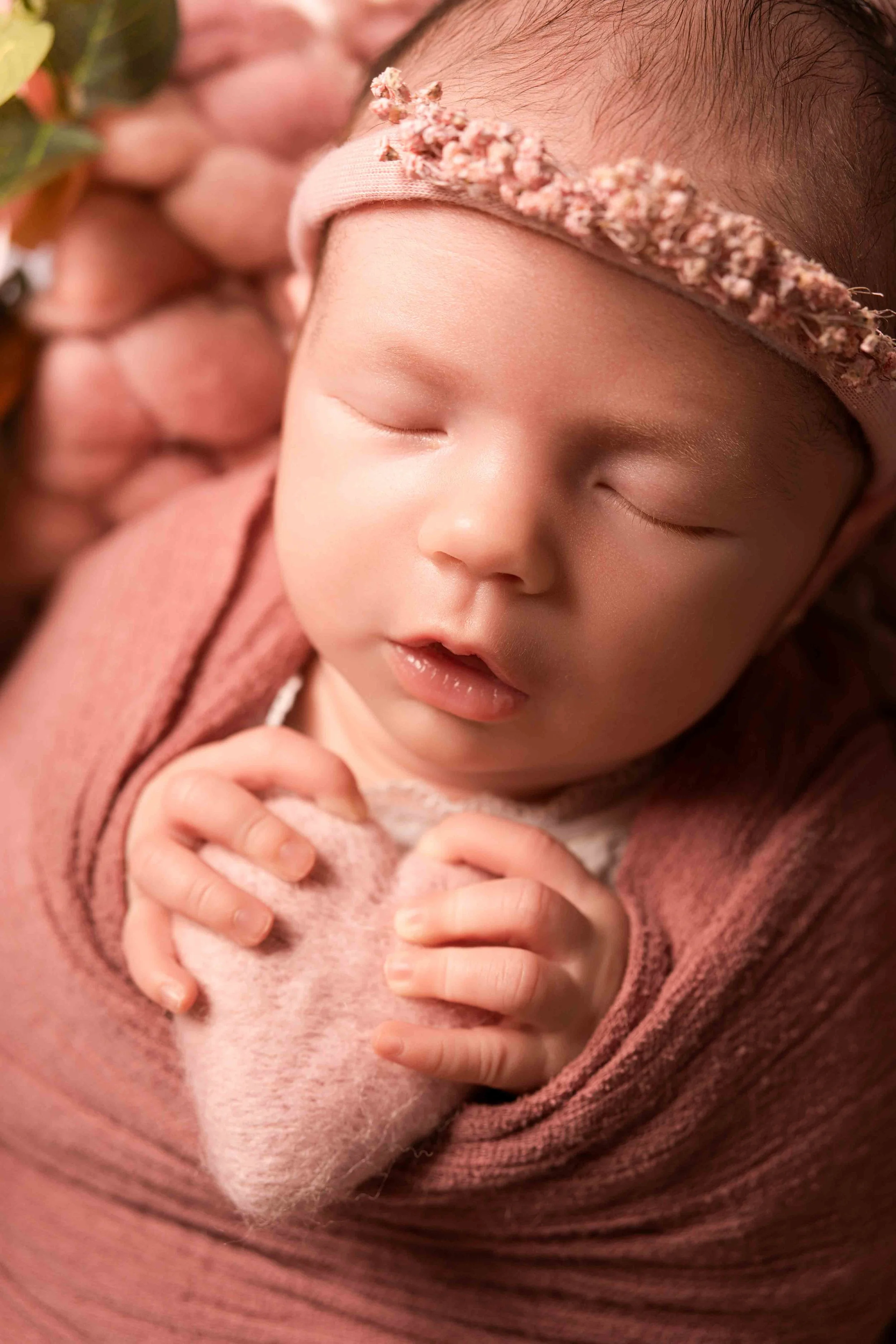 Ein neugeborenes Baby schläft friedlich, getragen in den Armen eines Erwachsenen, mit einem pinken Haarband mit kleinen Blumen und einem pinken Strickpullover.
