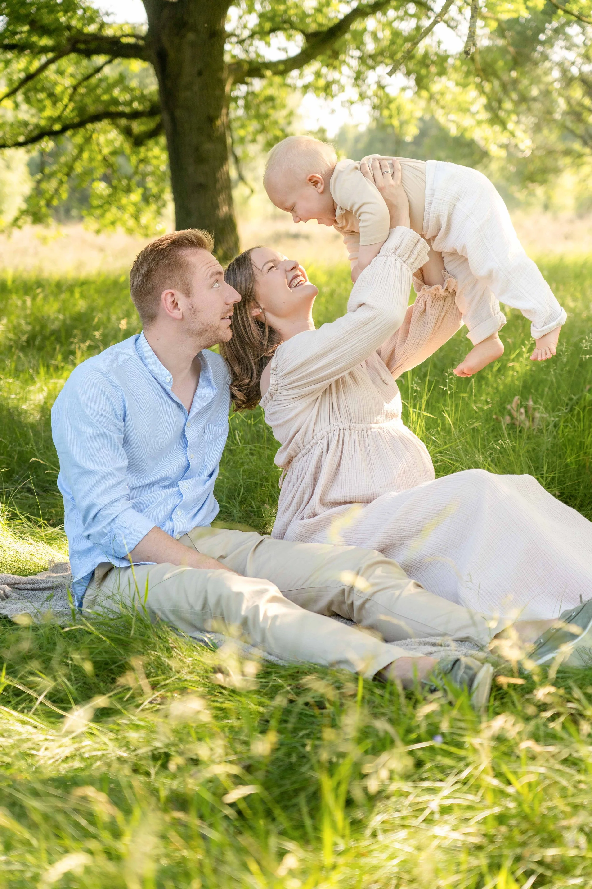 Familie besteht aus einem Mann, einer Frau und einem Baby, die im Freien unter einem Baum auf einer Wiese sitzen. Die Frau hält das Baby in die Luft, während der Mann und die Frau lachen und den Moment genießen.