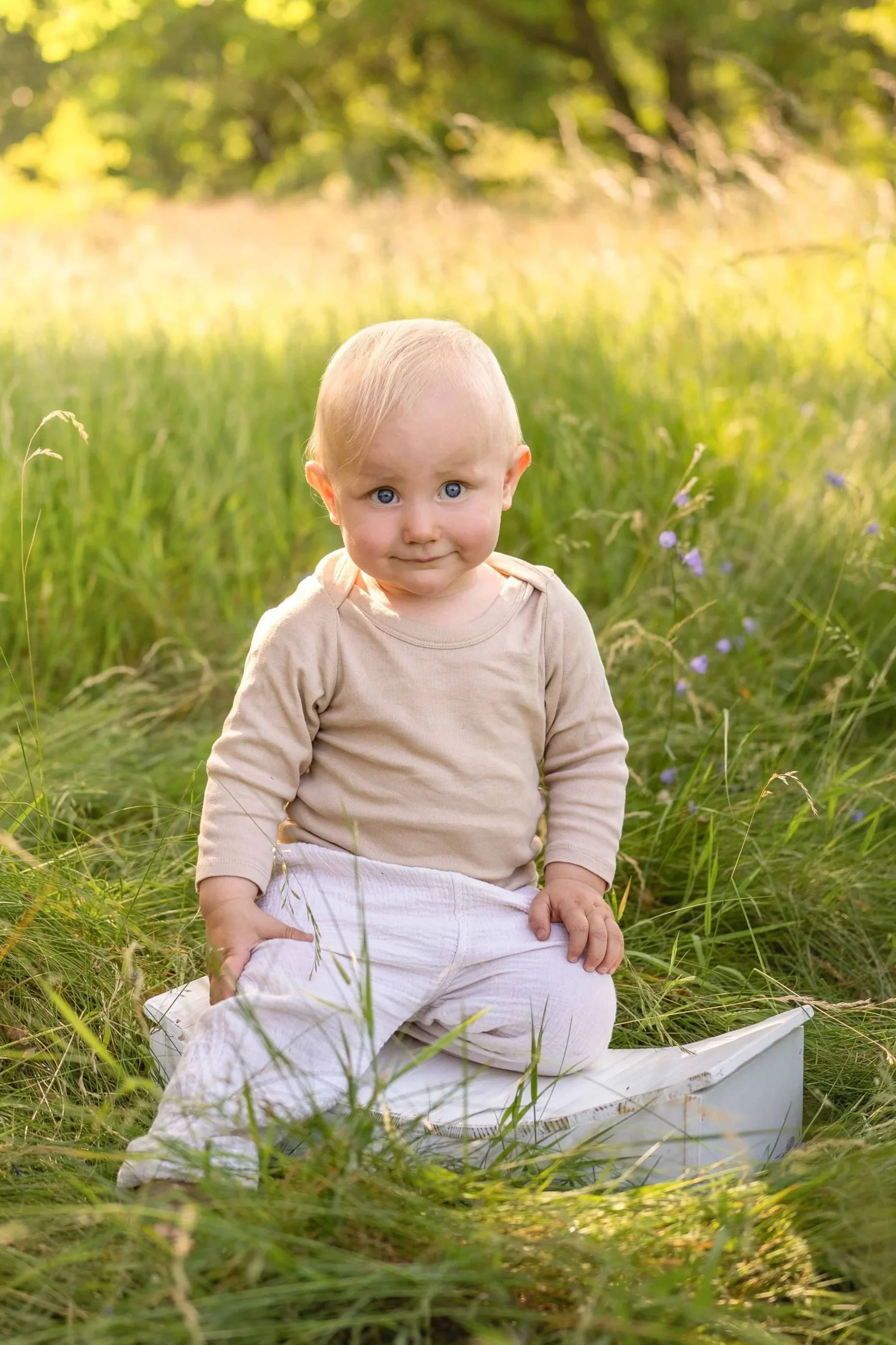 Kleines Kind mit blonden Haaren und blauen Augen sitzt im Gras im Freien, umgeben von grünen Pflanzen und Blumen, bei Sonnenuntergang.