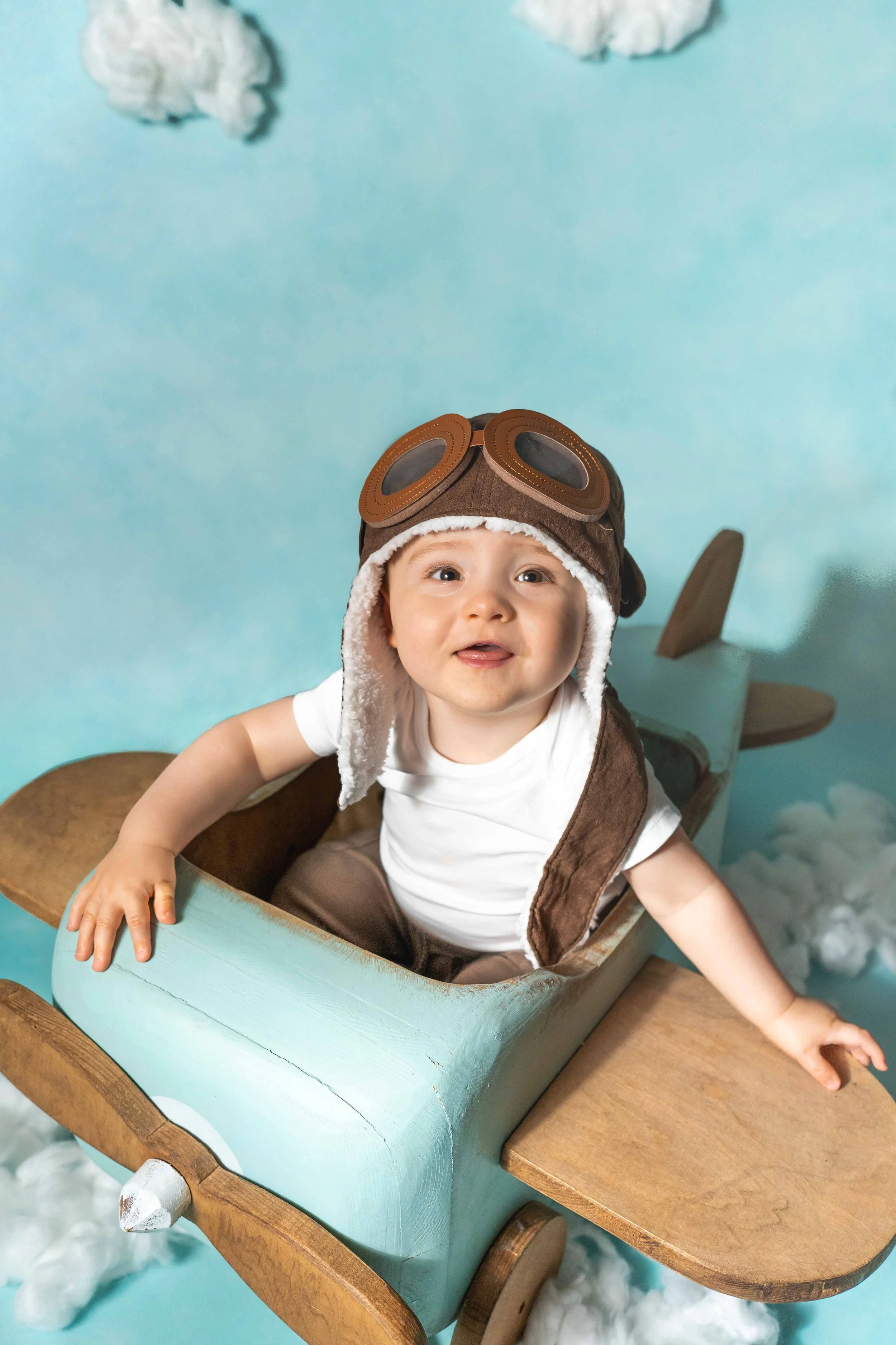 Süßer Baby in Flugzeug-Kostüm mit Pilotenhelm und Brille, sitzend auf einem selbstgebauten Holzflugzeug, mit Himmel- und Wolkendesign im Hintergrund.