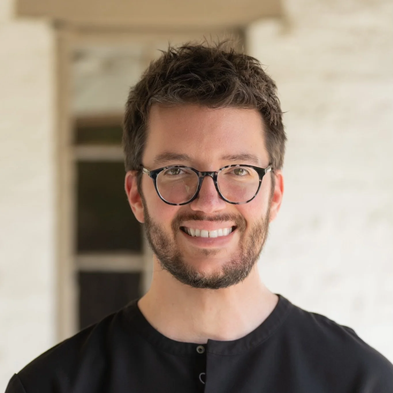 A smiling man with glasses and a beard wearing a black shirt indoors.