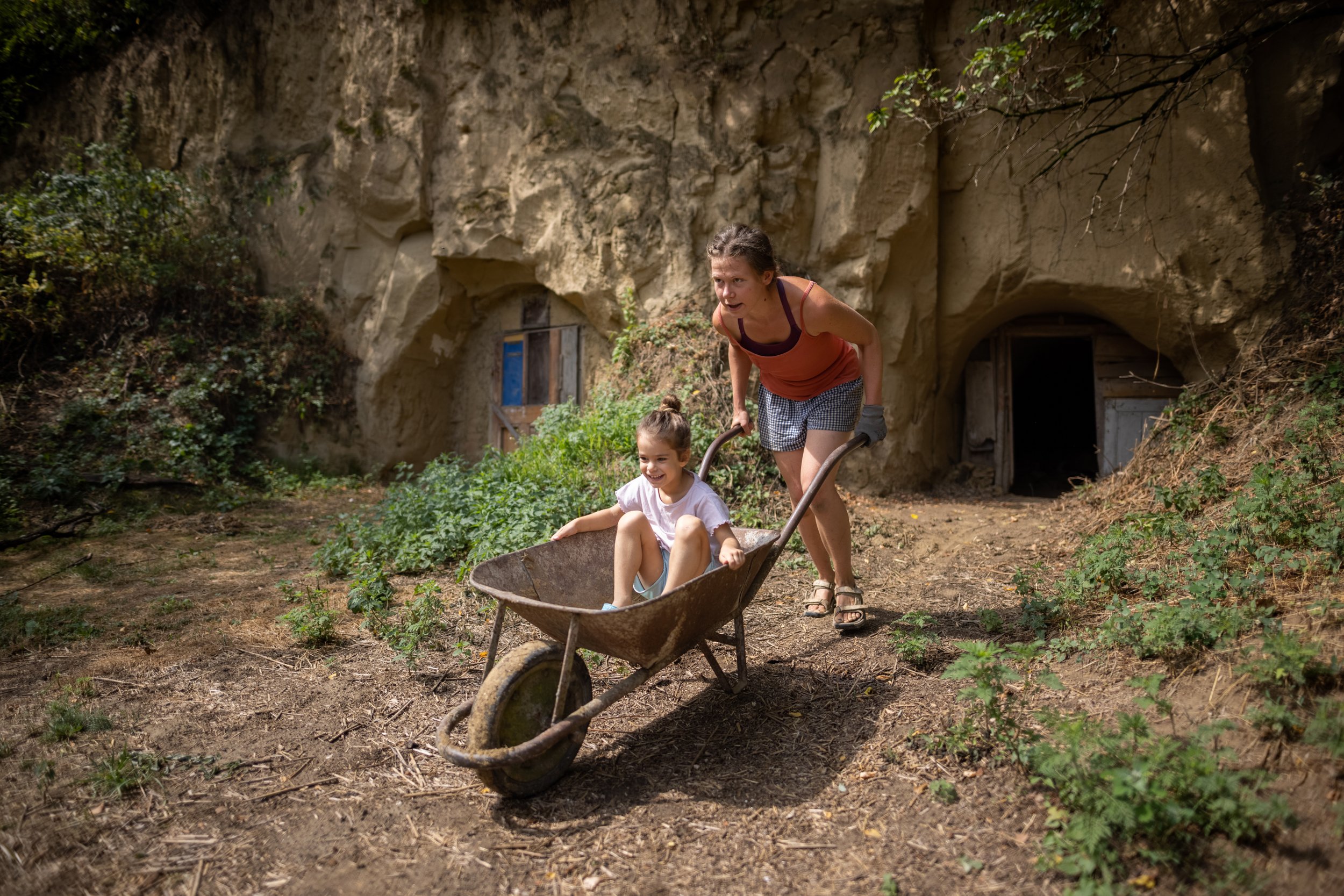  Virág pushing a little girl in a wheelbarrow in Nagyszékely on August 9, 2022. Even amid a tiring weekday – in this case, the renovation of an adobe building – you can still have fun. The cellars carved into the loess wall in the background are also