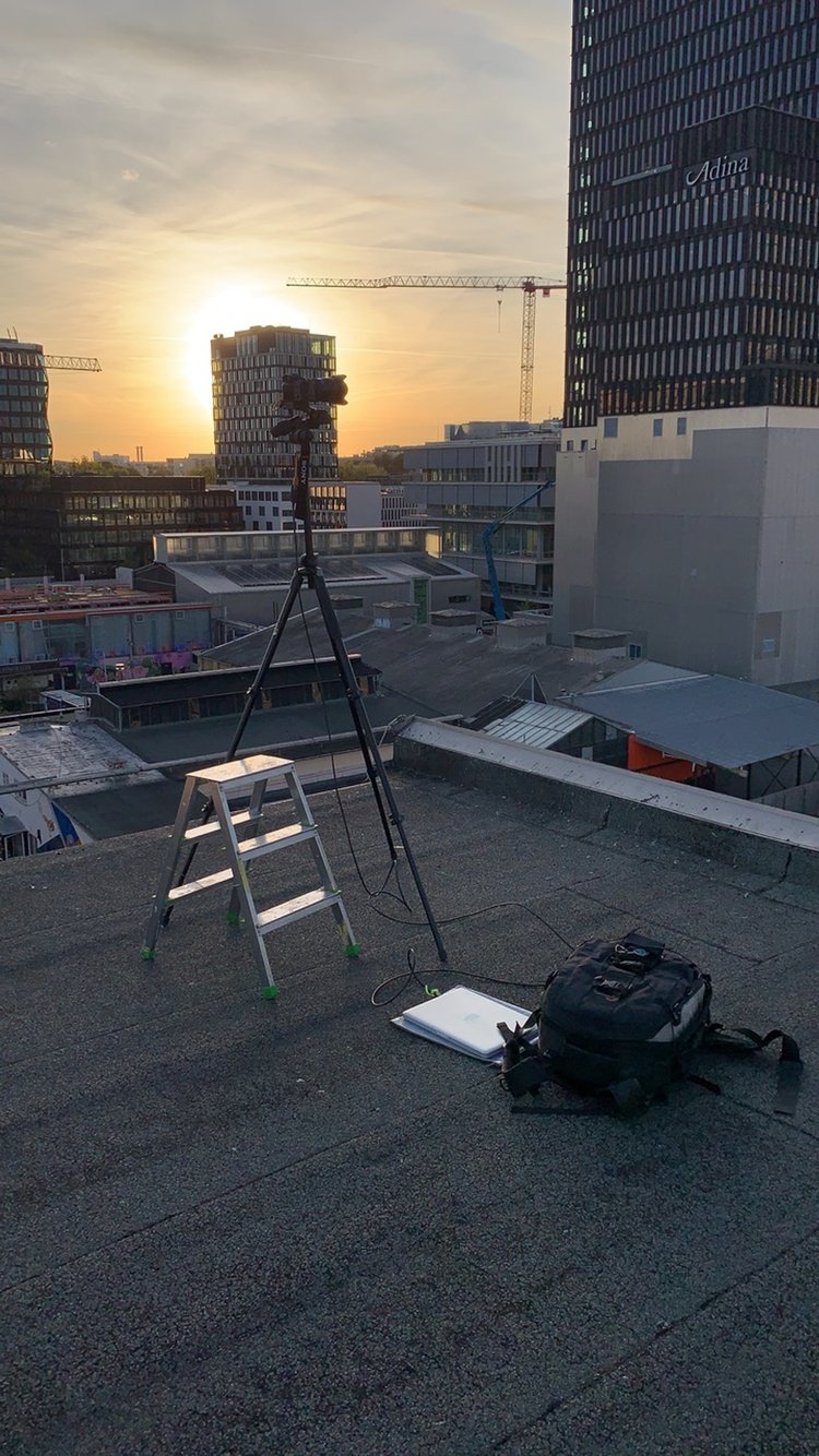 Sonnenuntergang auf einem Dach mit Kameraausrüstung, Stativ, Laptop und Rucksack. Im Hintergrund sind Kräne und moderne Gebäude zu sehen.