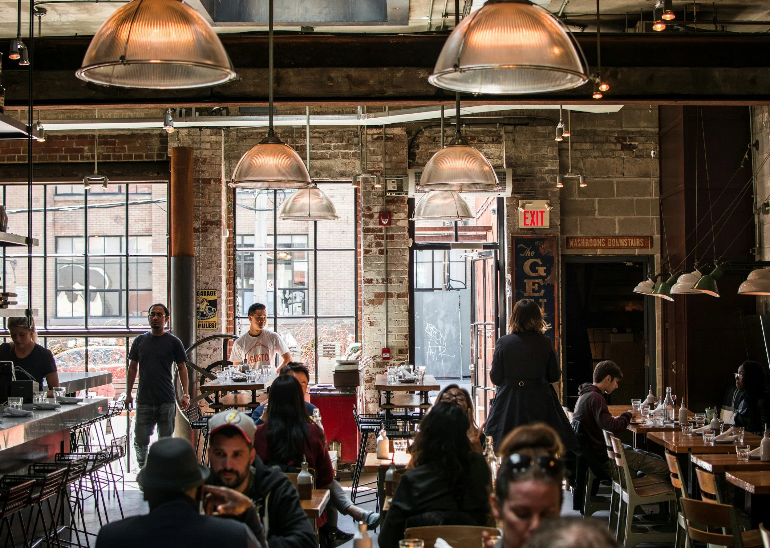 Interior of a busy restaurant or cafe with large windows, exposed brick walls, hanging pendant lights, and people dining and standing, some near the open door.