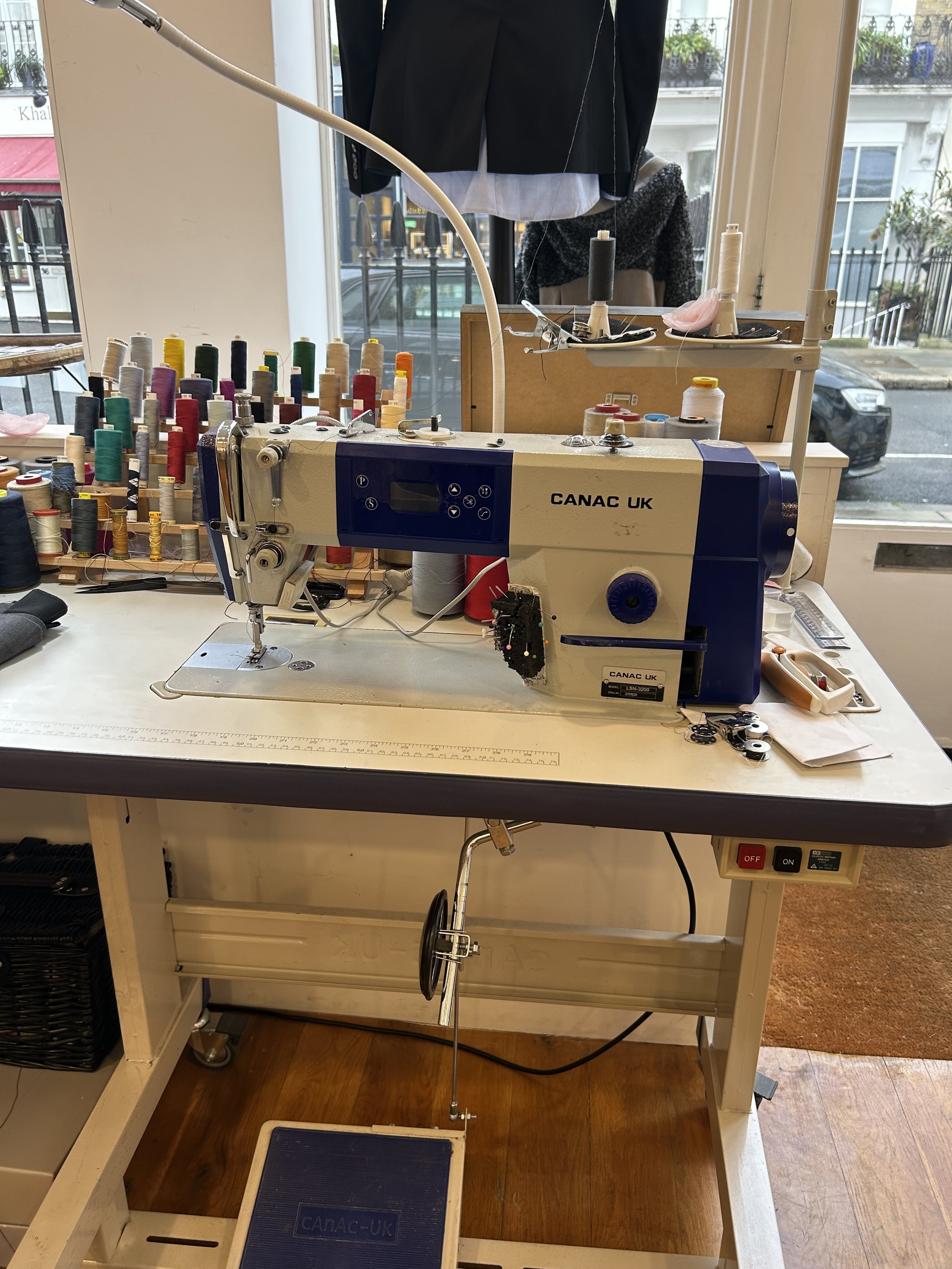 A sewing machine on a work table surrounded by spools of thread, with fabric and sewing accessories, in a sewing or tailoring shop.