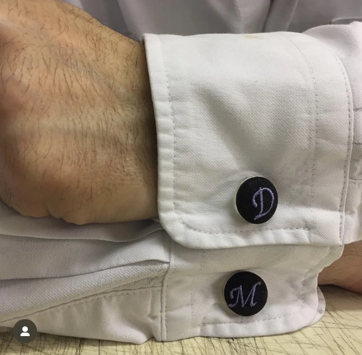 A close-up of a person's wrist with visible arm hair, wearing a white shirt with black embroidered buttons labeled 'D' and 'M'.