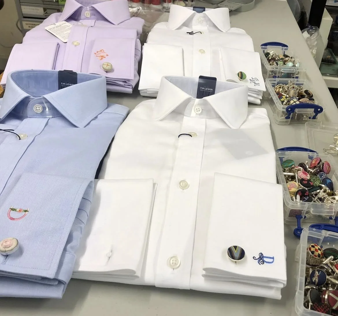 Several folded dress shirts in light purple, white, and pale blue colors displayed on a table, with small decorative pins and cufflinks placed on some shirts and in small plastic containers on the side.