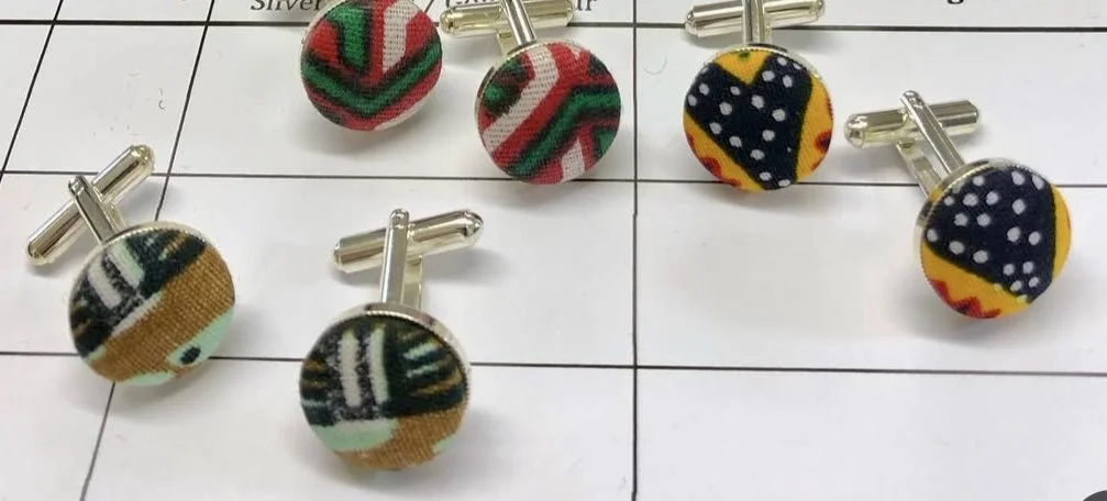 Five fabric-covered cufflinks with colorful, patterned designs placed on a white grid paper background.