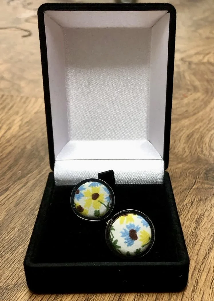 A pair of cufflinks with a daisy flower pattern in a black jewelry box on a wooden surface.