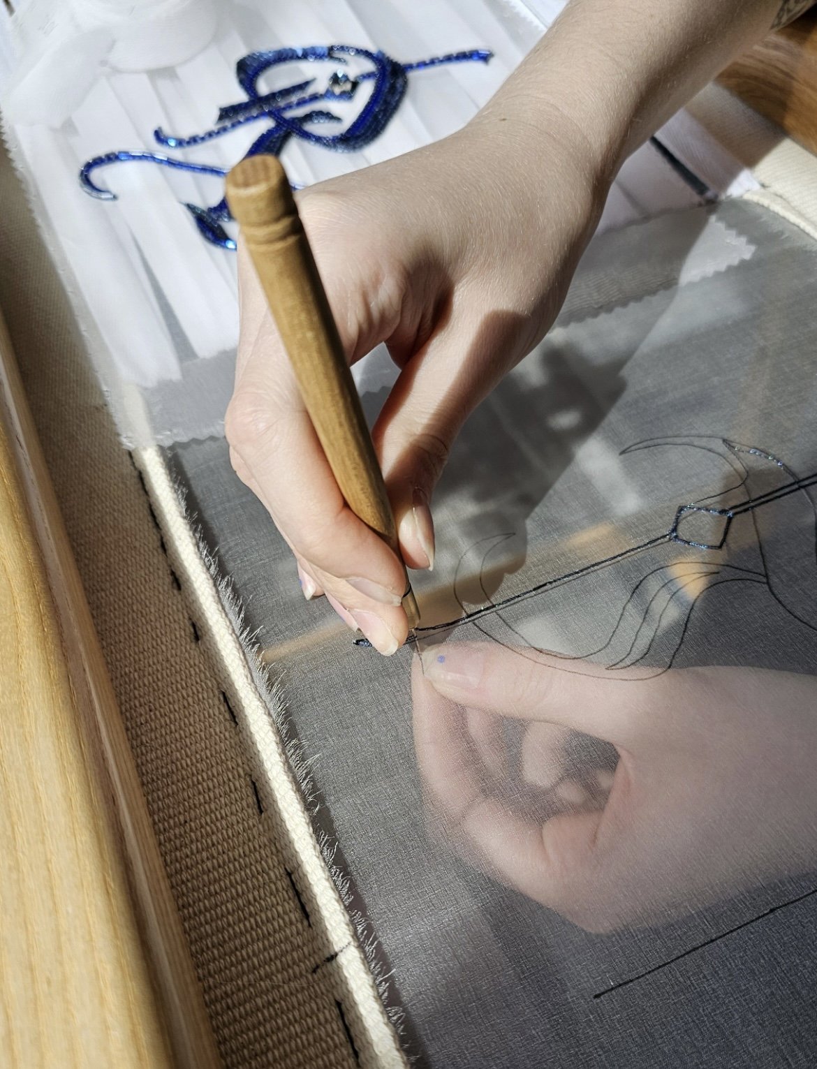 Close-up of a person's hand holding a stylus and working on a paper pattern for embroidery or sewing on a clear fabric, with embroidered design visible in the background.