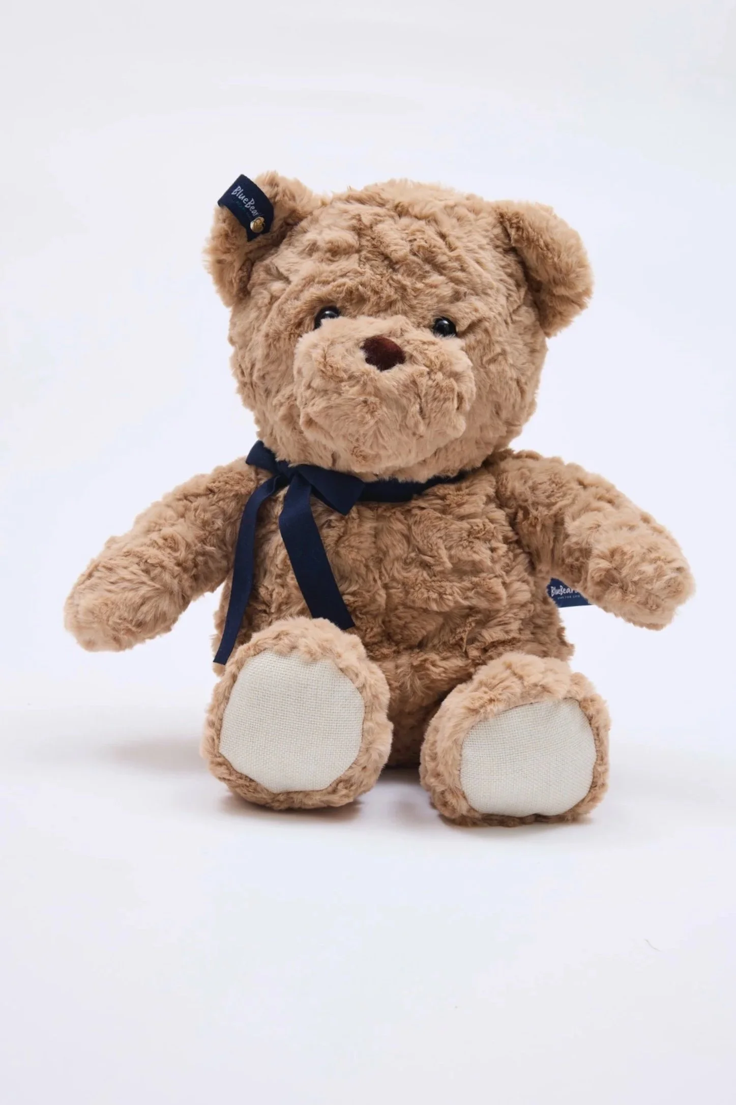 A plush teddy bear with light brown fur, a dark blue ribbon around its neck, and a matching dark blue tag on its ear, sitting against a white background.