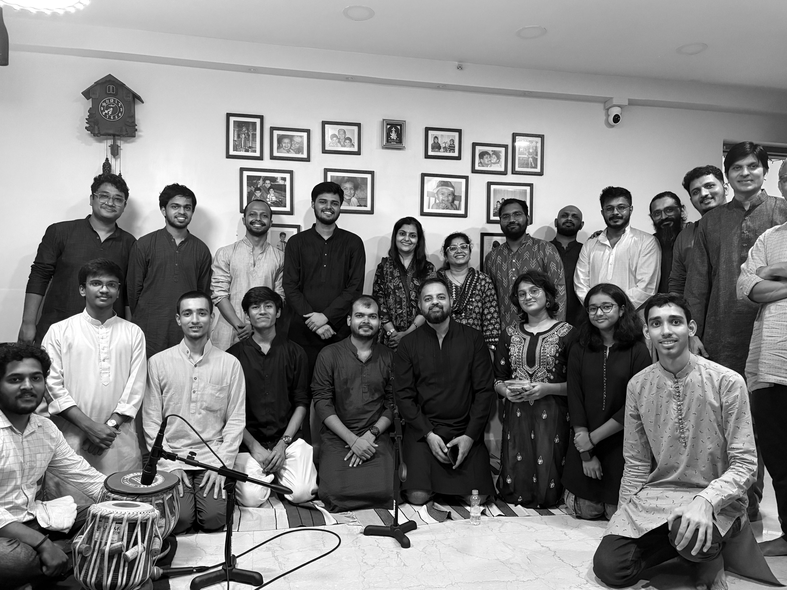 Group photo of people in traditional and casual attire, sitting and standing in a room with framed pictures on the wall, a clock, and musical instruments.